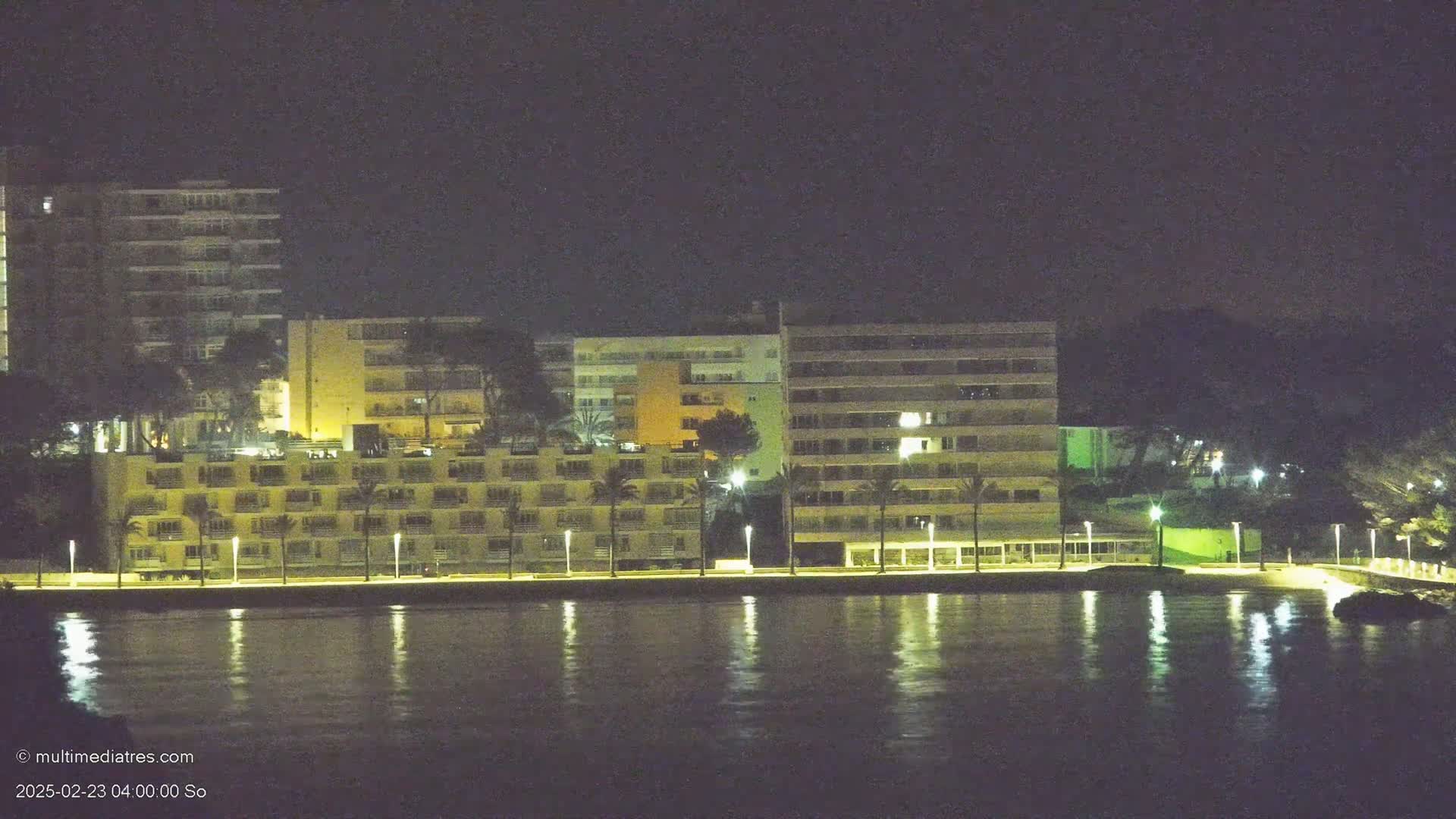 A row of illuminated buildings line a waterfront at night, their lights reflecting on calm water.