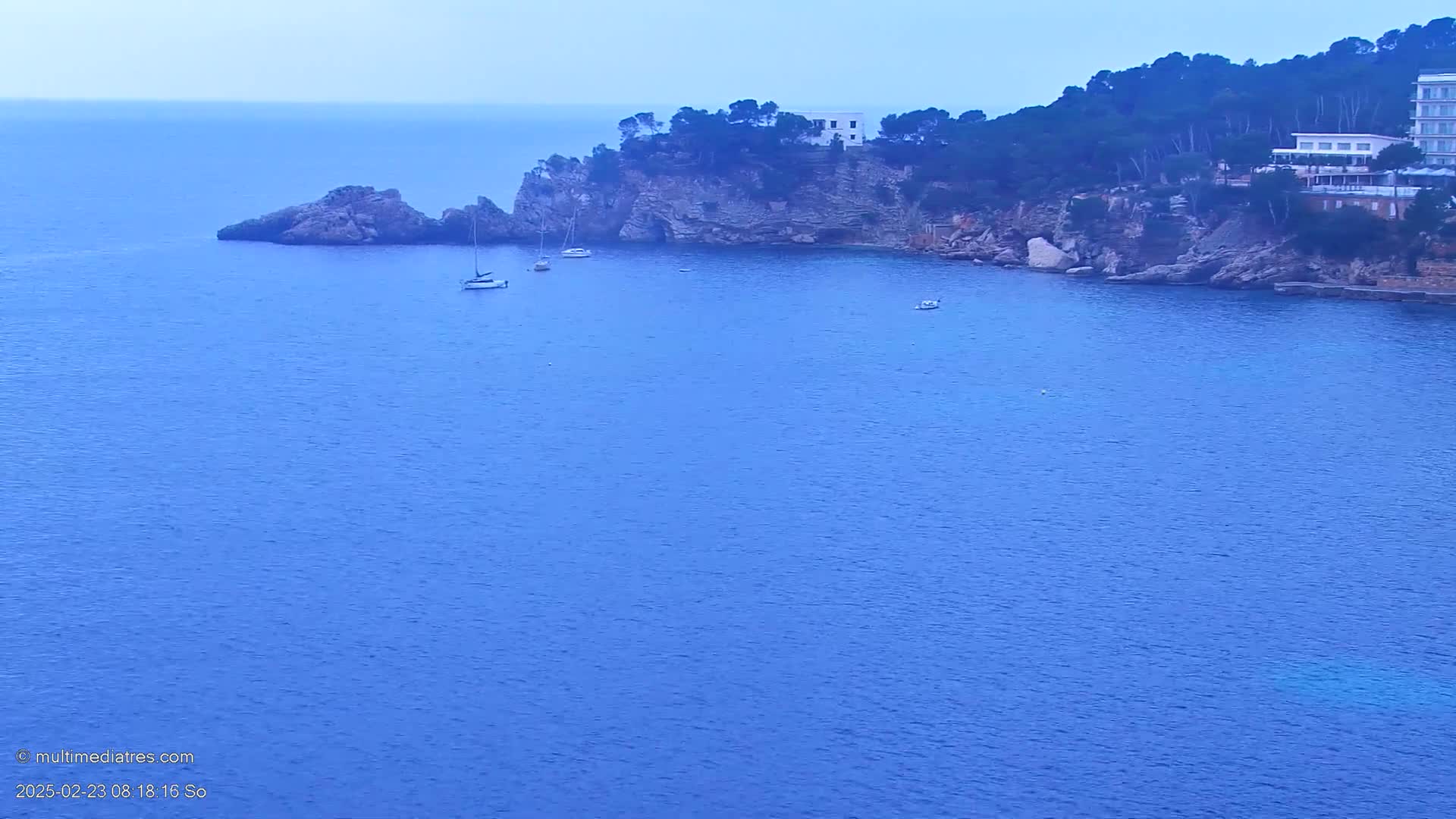 A tranquil, blue sea laps against a rocky coastline dotted with a few boats and buildings under an overcast sky.