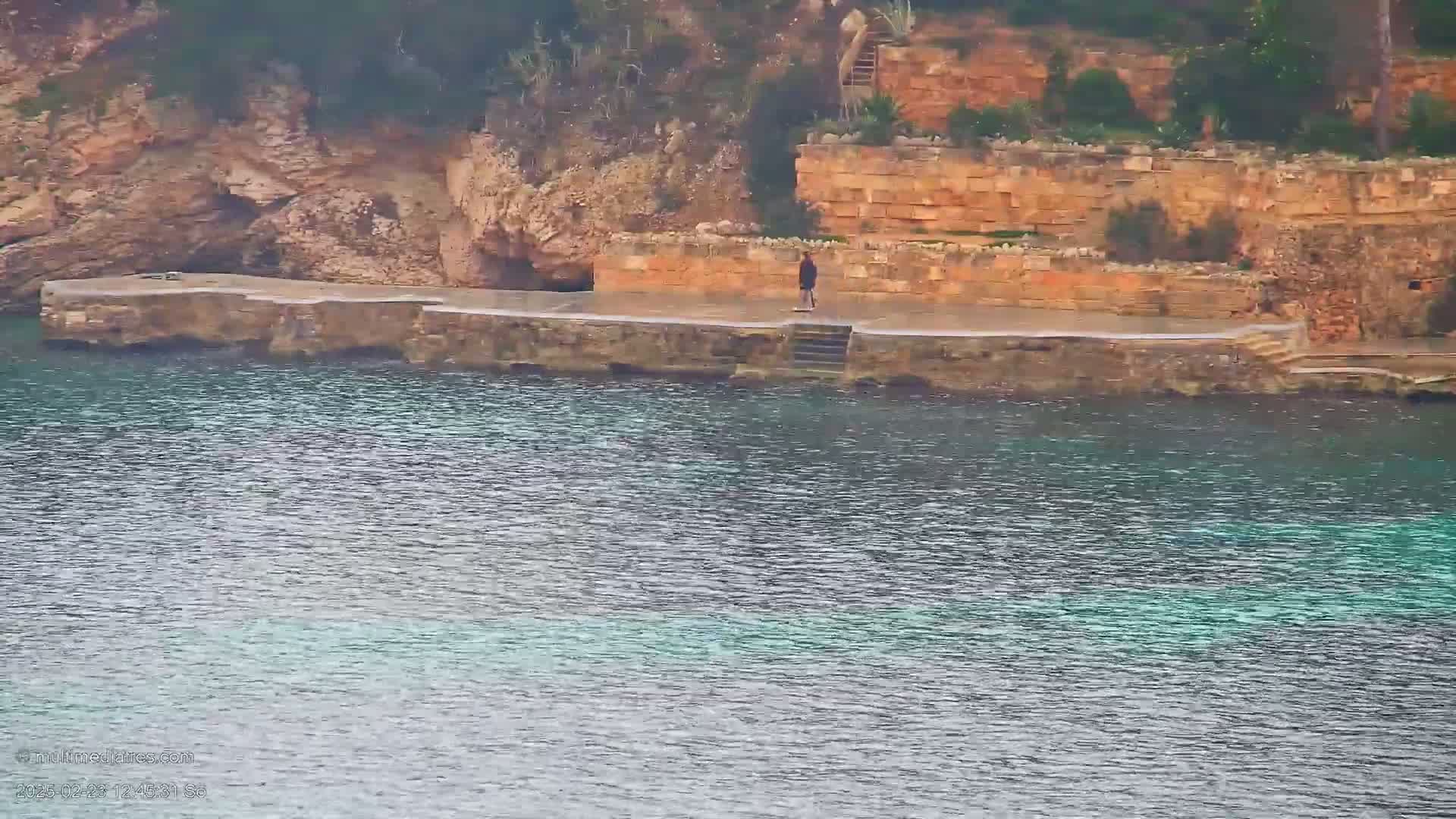 A person stands on a stone platform next to calm, teal water, backed by a rocky cliff face with stonework.
