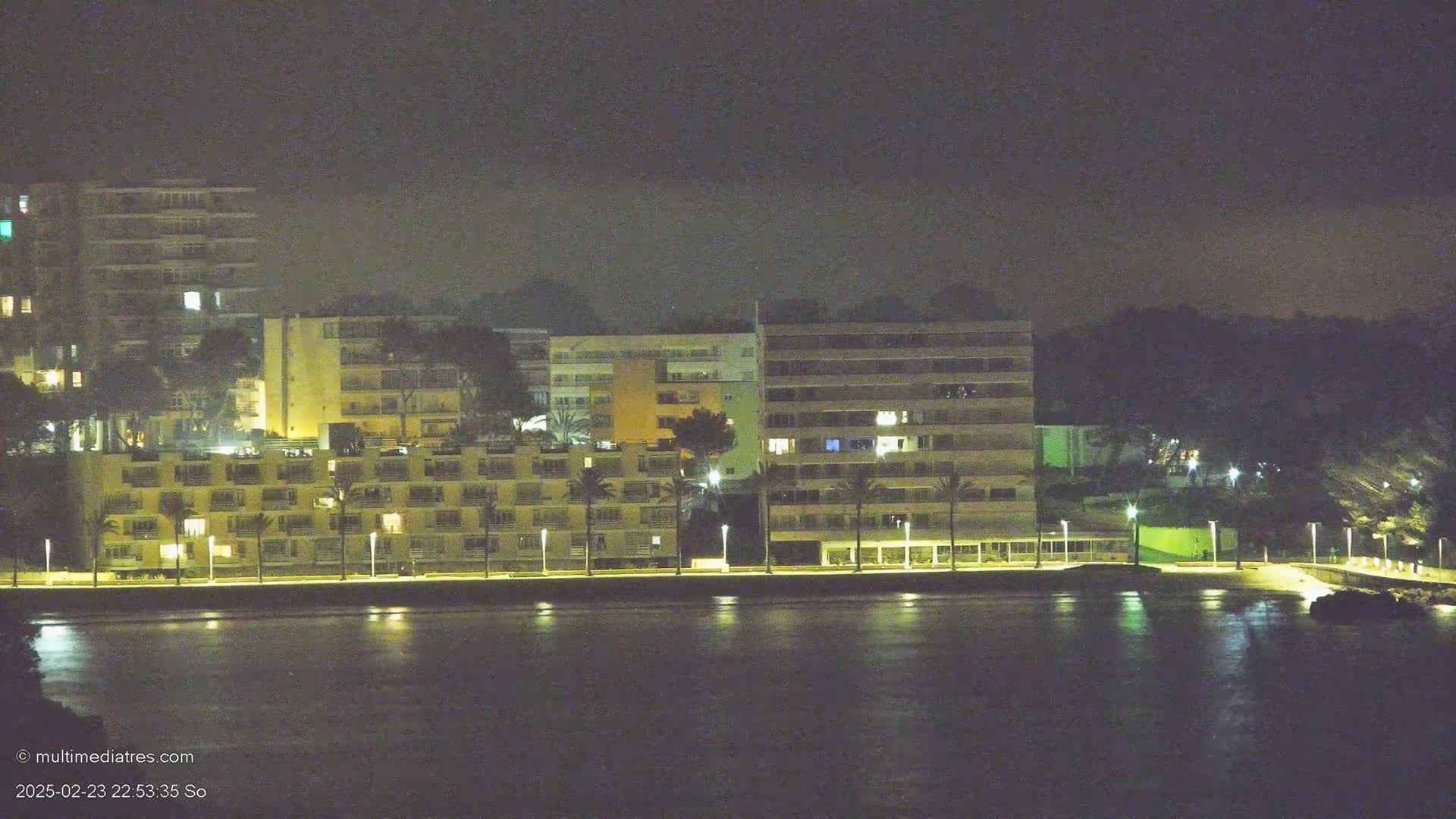 A nighttime view of a waterfront with several illuminated multi-story buildings and palm trees lining a paved walkway, all under a dark, hazy sky.
