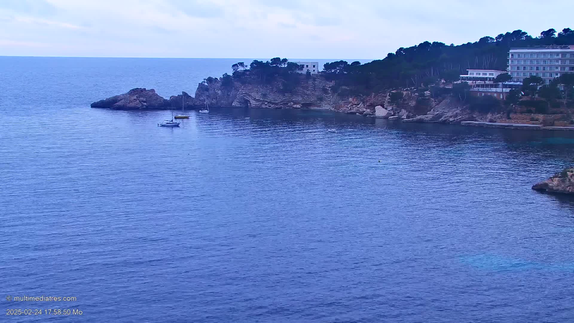 A calm, blue sea laps against a rocky coastline dotted with a few small boats and a large hotel nestled among the trees under an overcast sky.