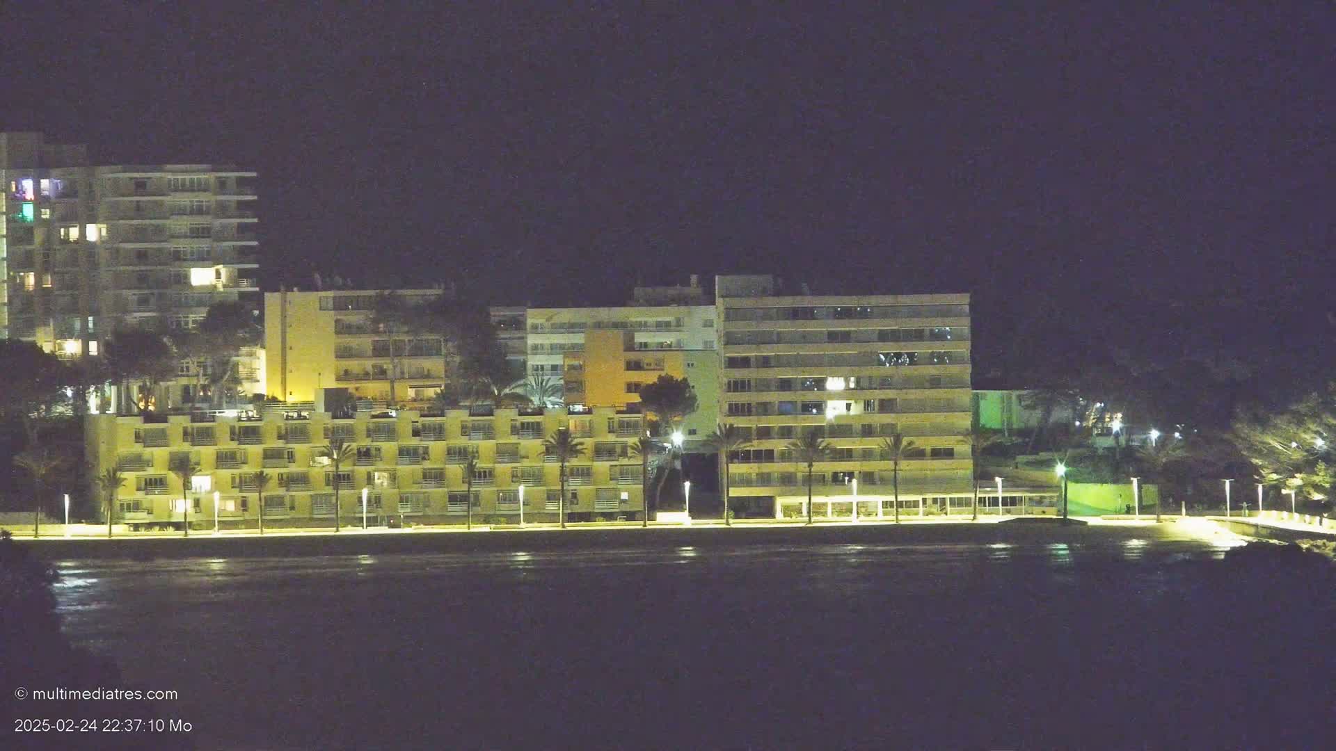A row of lit apartment buildings and hotels lines a dark waterfront at night.