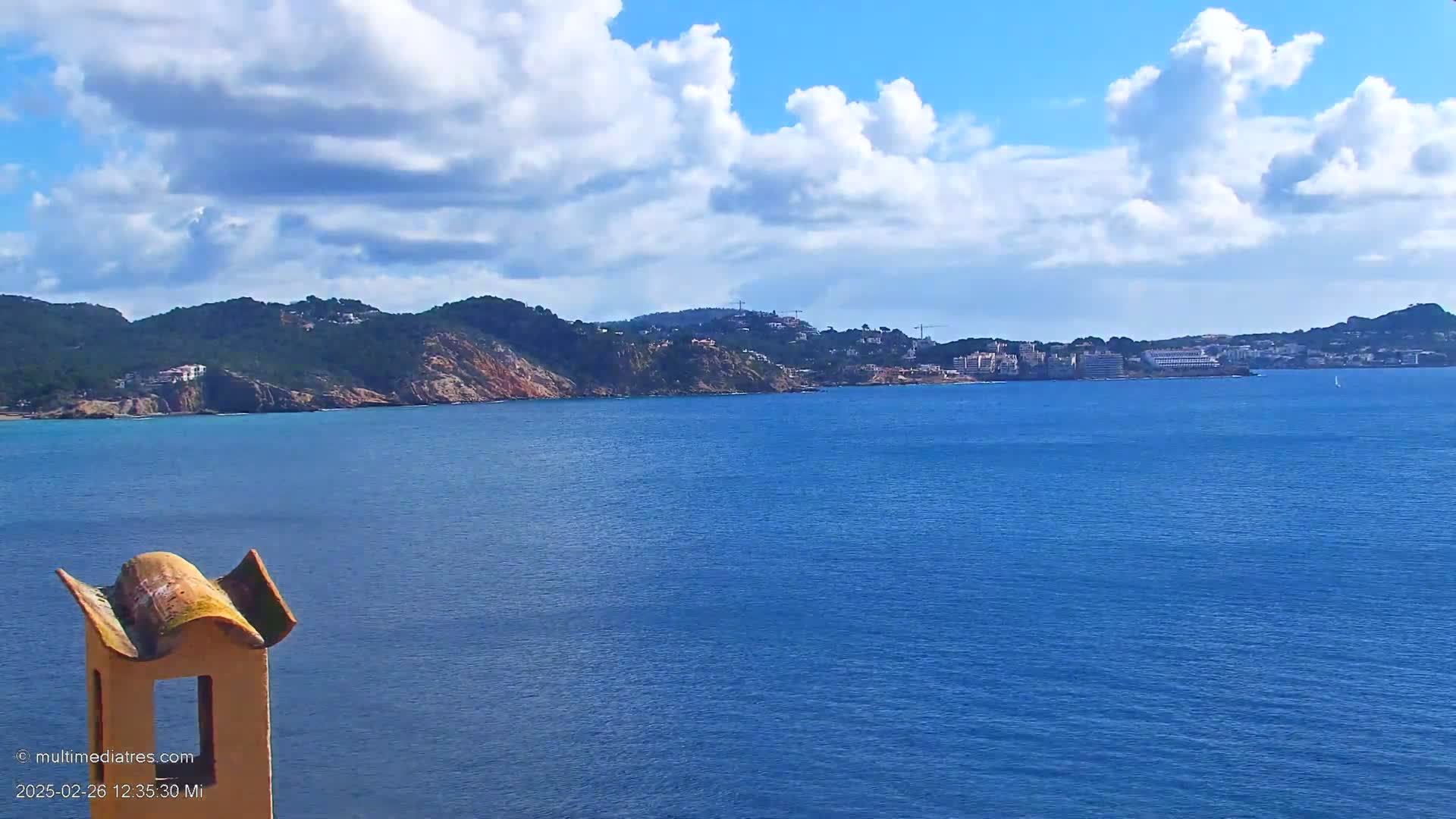 A partly cloudy day reveals a coastal scene with a blue ocean, a rocky hillside with buildings, and a portion of a unique yellow chimney in the foreground.
