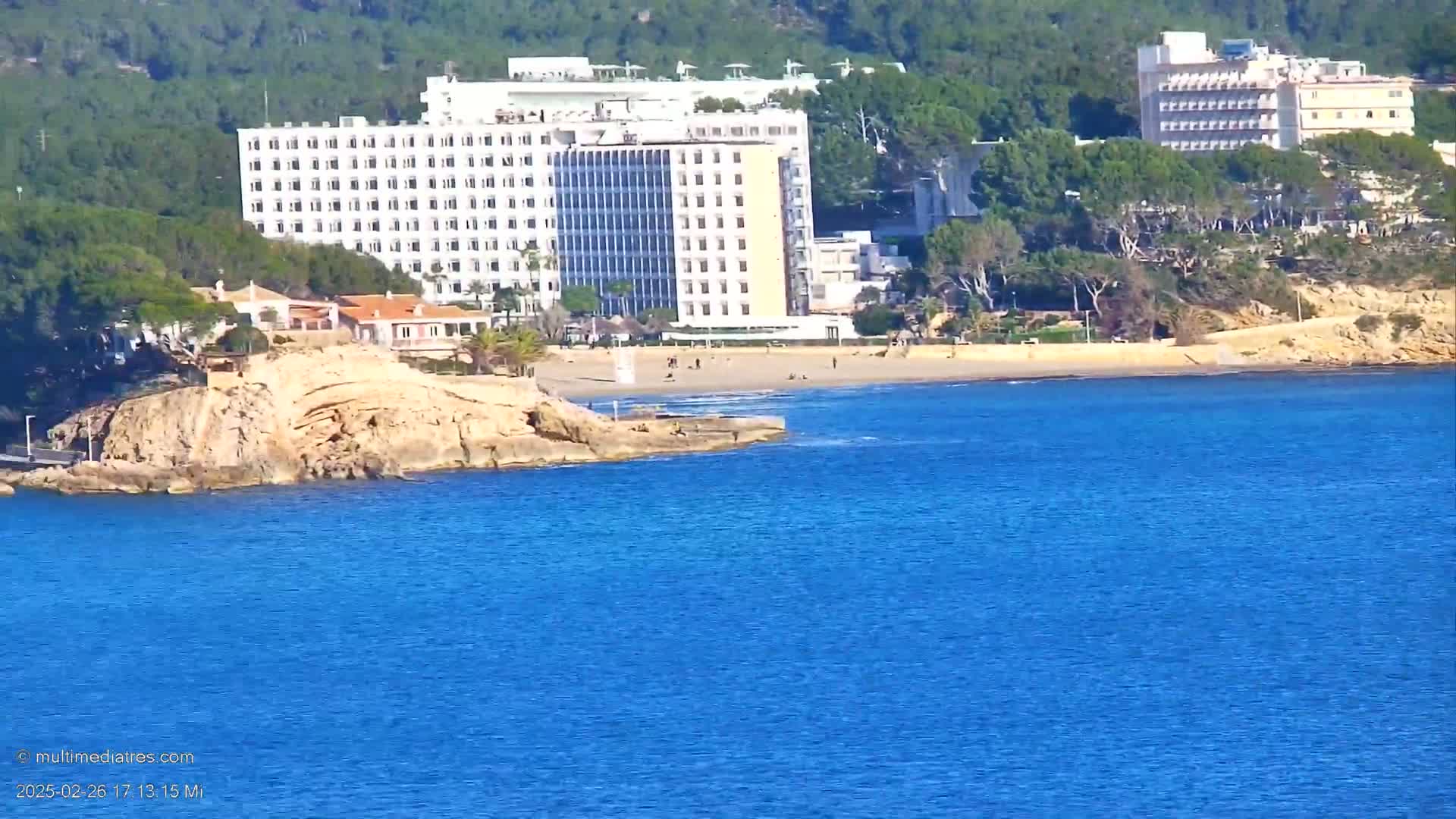 A sandy beach with a few people on it sits below a large white hotel and other buildings nestled into a green hillside overlooking a calm blue sea.
