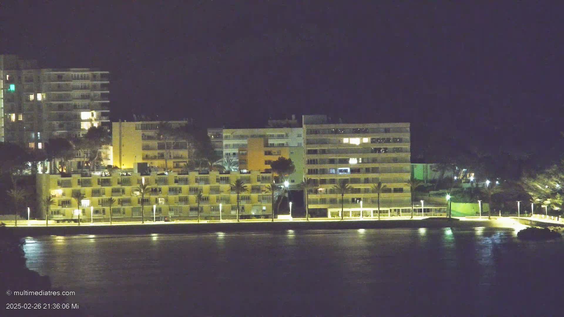 Several multi-story buildings with illuminated windows are situated along a waterfront at night, with calm water reflecting the lights.