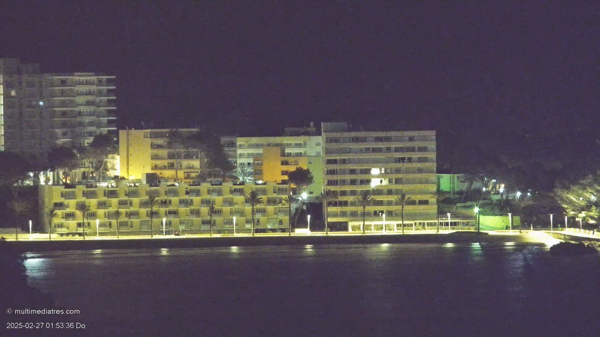 A row of brightly lit multi-story buildings stands along a dark waterfront at night.