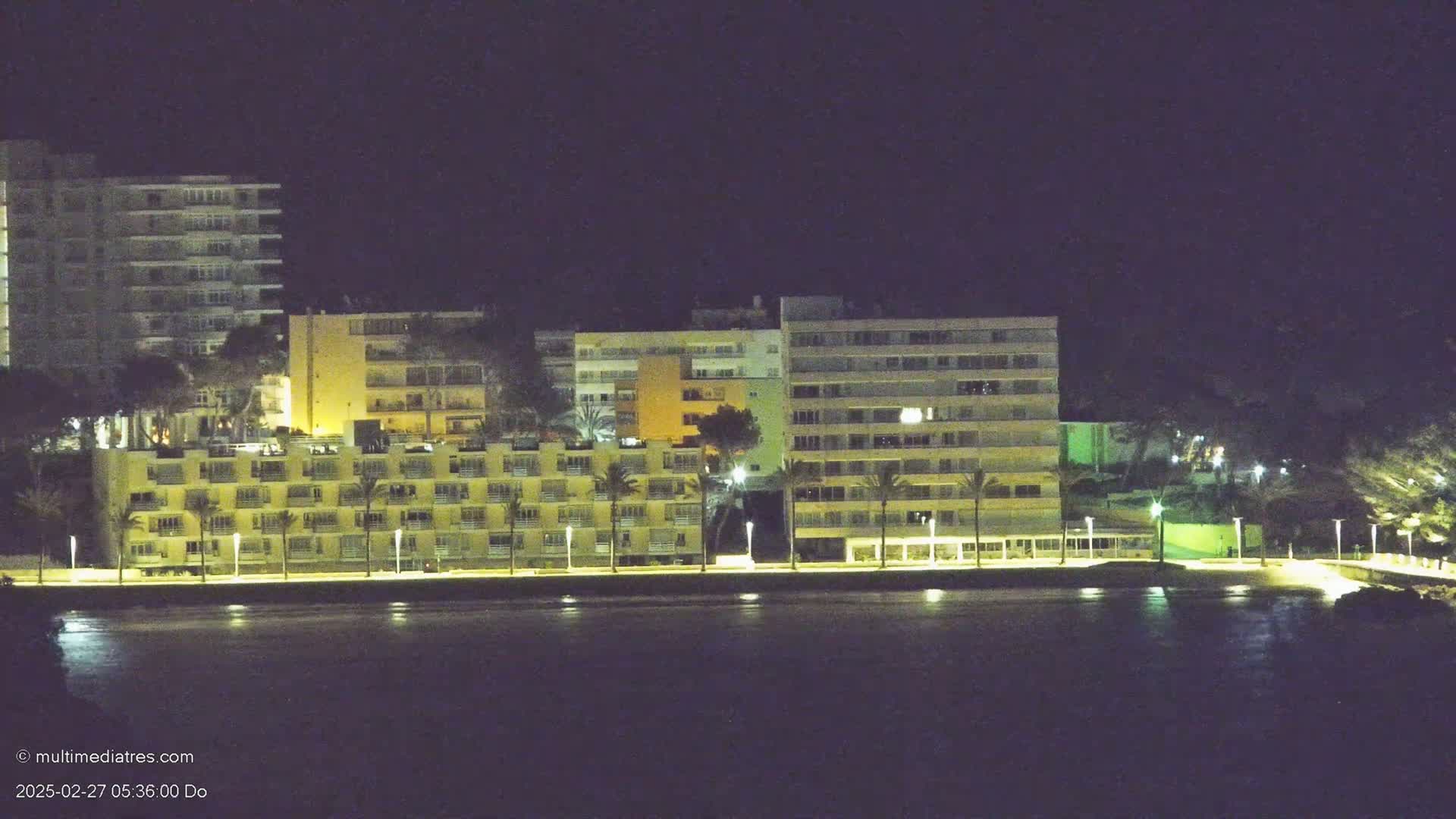 A row of illuminated beachfront buildings stands at night next to a dark body of water.