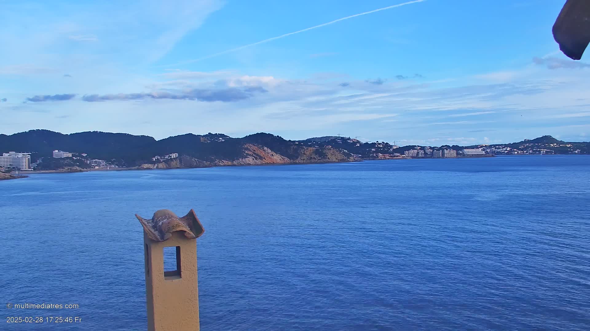A calm blue sea stretches out before a hilly coastline with buildings, under a mostly clear sky with a few clouds.