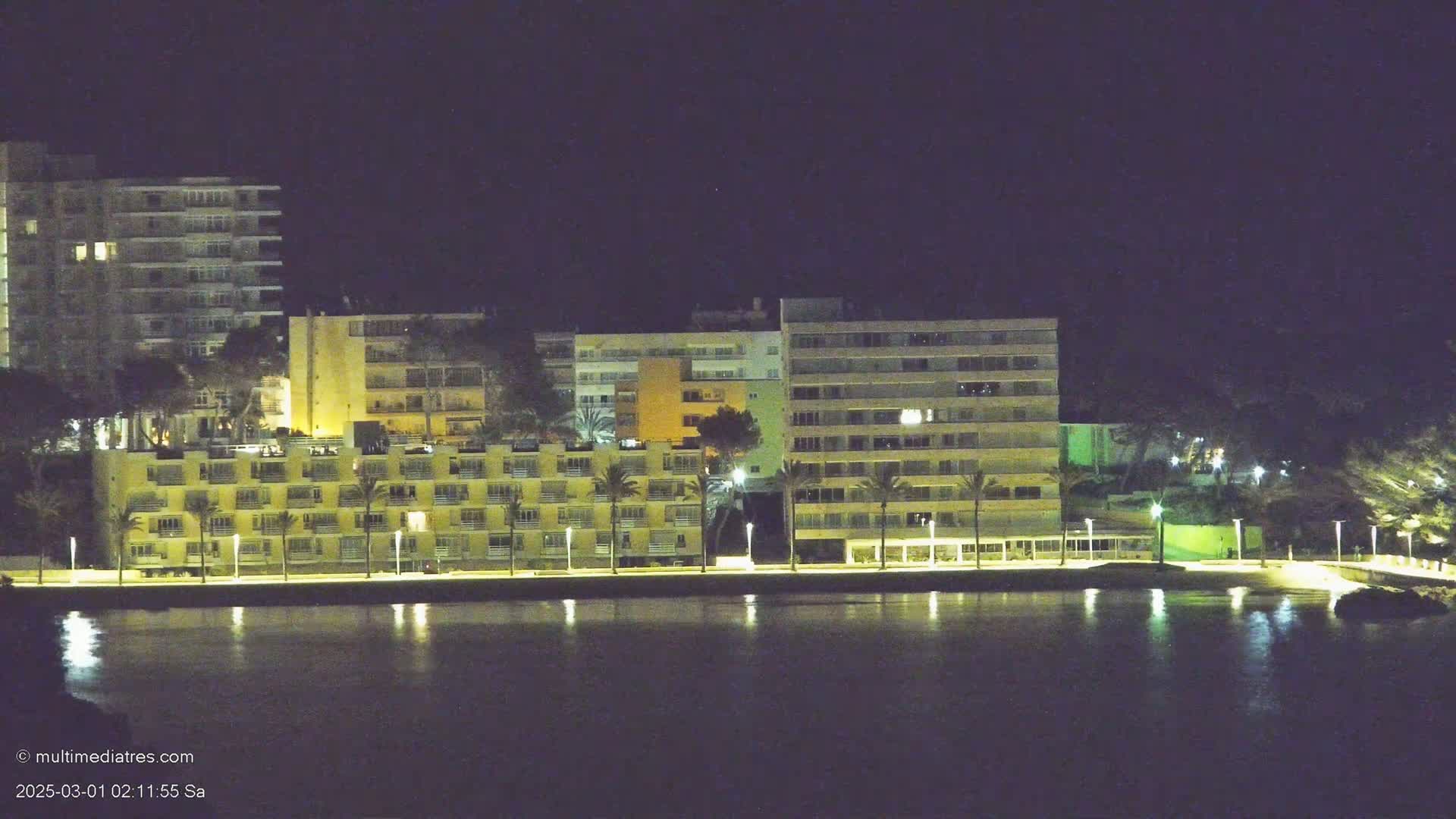 A row of illuminated buildings stands along a calm waterfront at night.