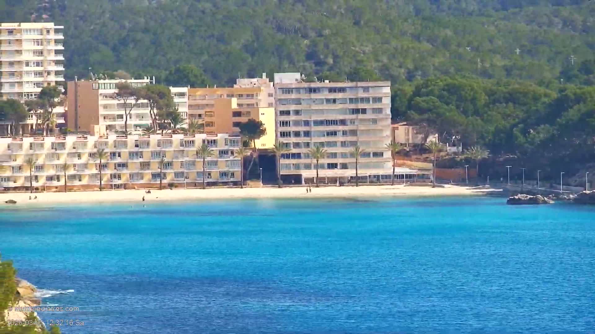 A sandy beach with several multi-story buildings sits on a bright sunny day next to a calm, vibrantly blue body of water.