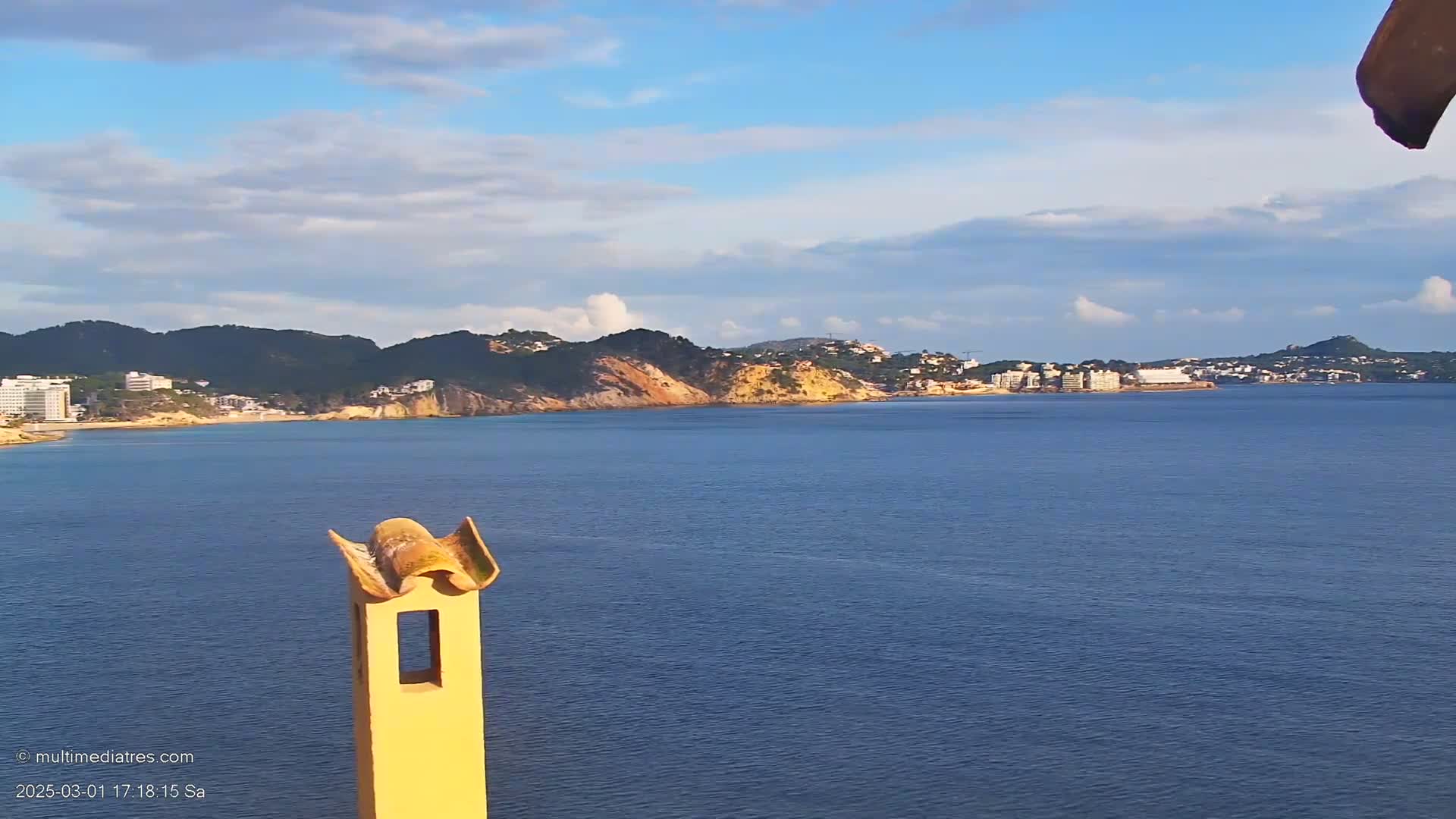 A partly cloudy day reveals a coastal scene of a calm blue sea, a sandy beach backed by low hills dotted with buildings, and a portion of a pale yellow chimney in the foreground.