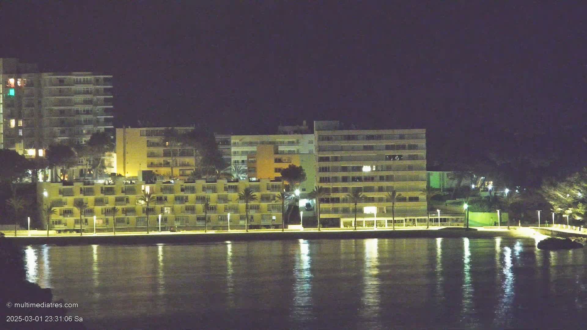 Illuminated buildings line a waterfront at night, their lights reflecting on calm water.