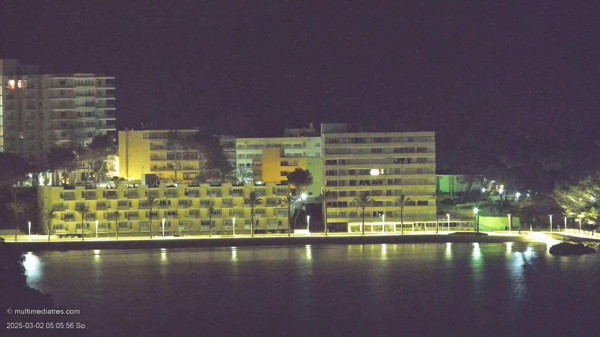 A row of illuminated apartment buildings sits along a calm body of water at night.