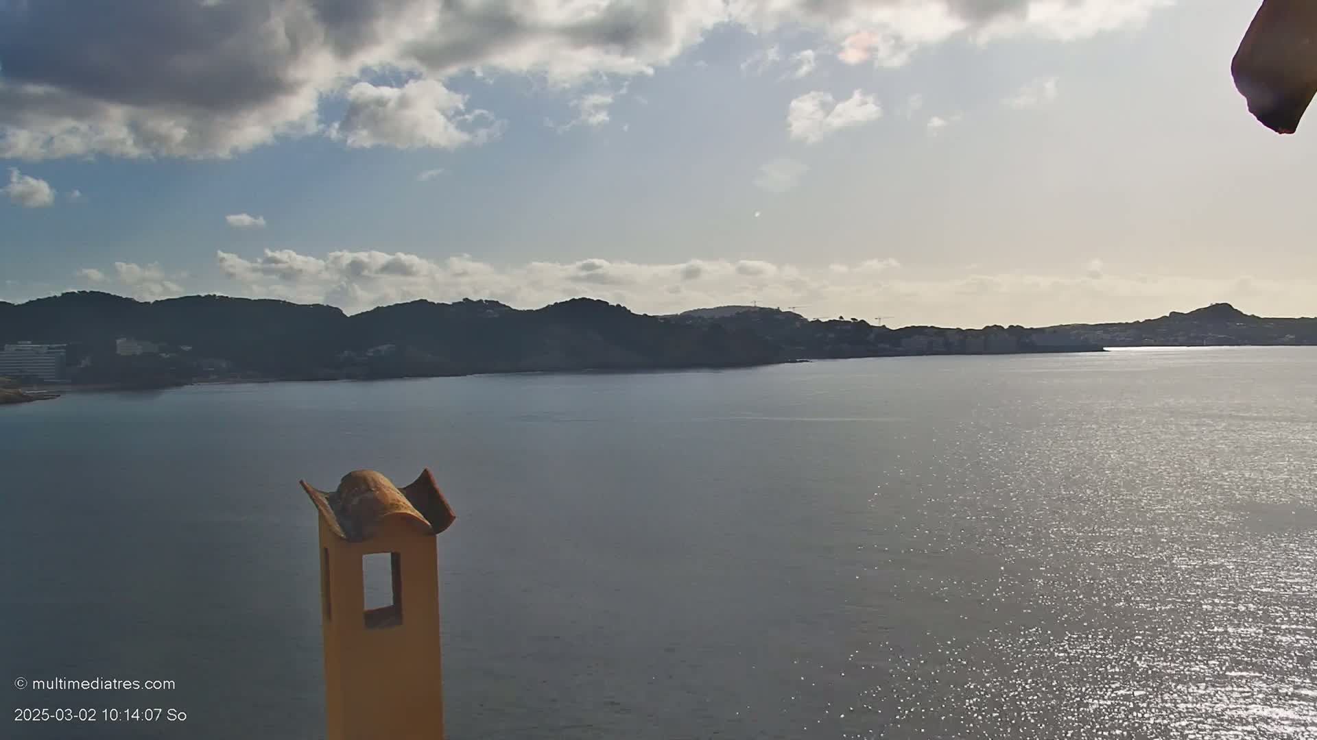 A partly cloudy day reveals a calm body of water reflecting sunlight, with dark hills and a coastline in the background, viewed from a vantage point near a yellow chimney.