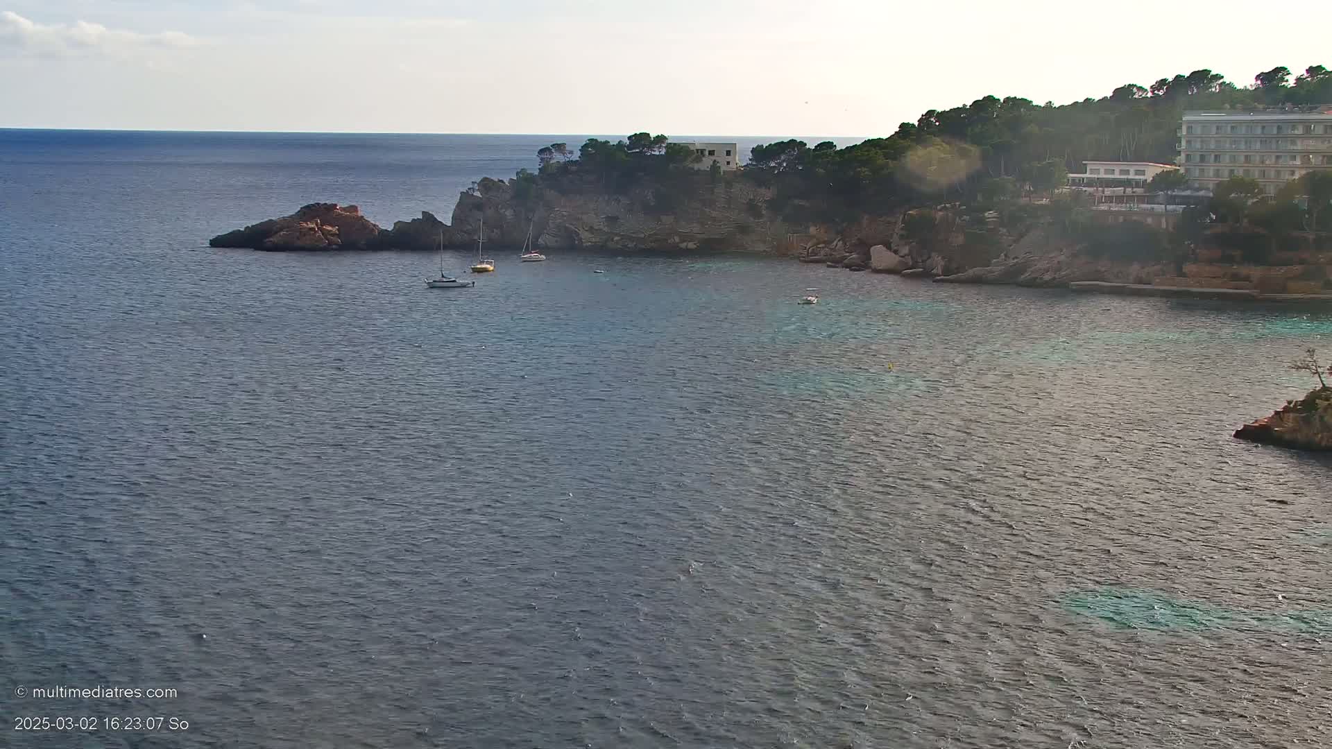 A calm sea laps against a rocky coastline where a few sailboats are anchored near a hotel.