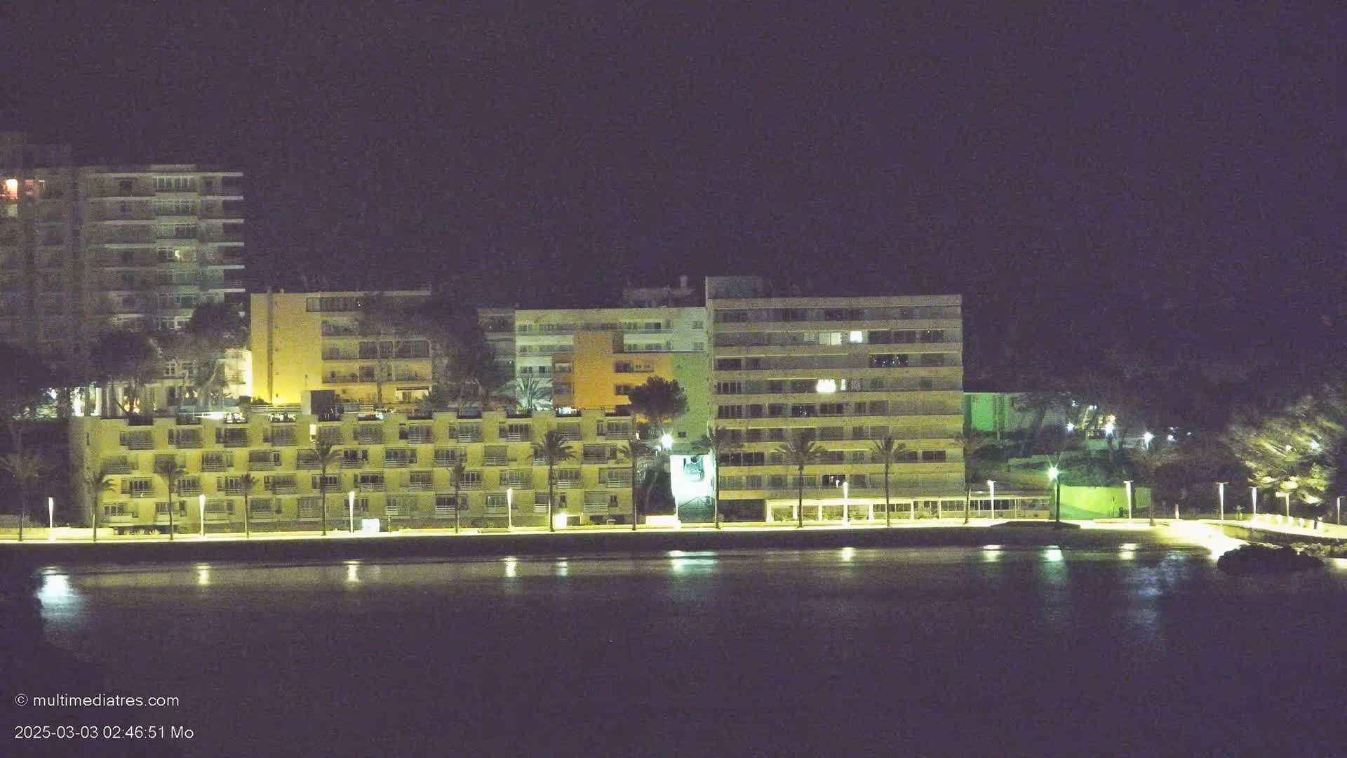 A row of illuminated multi-story buildings sits beside a dark body of water under a night sky.