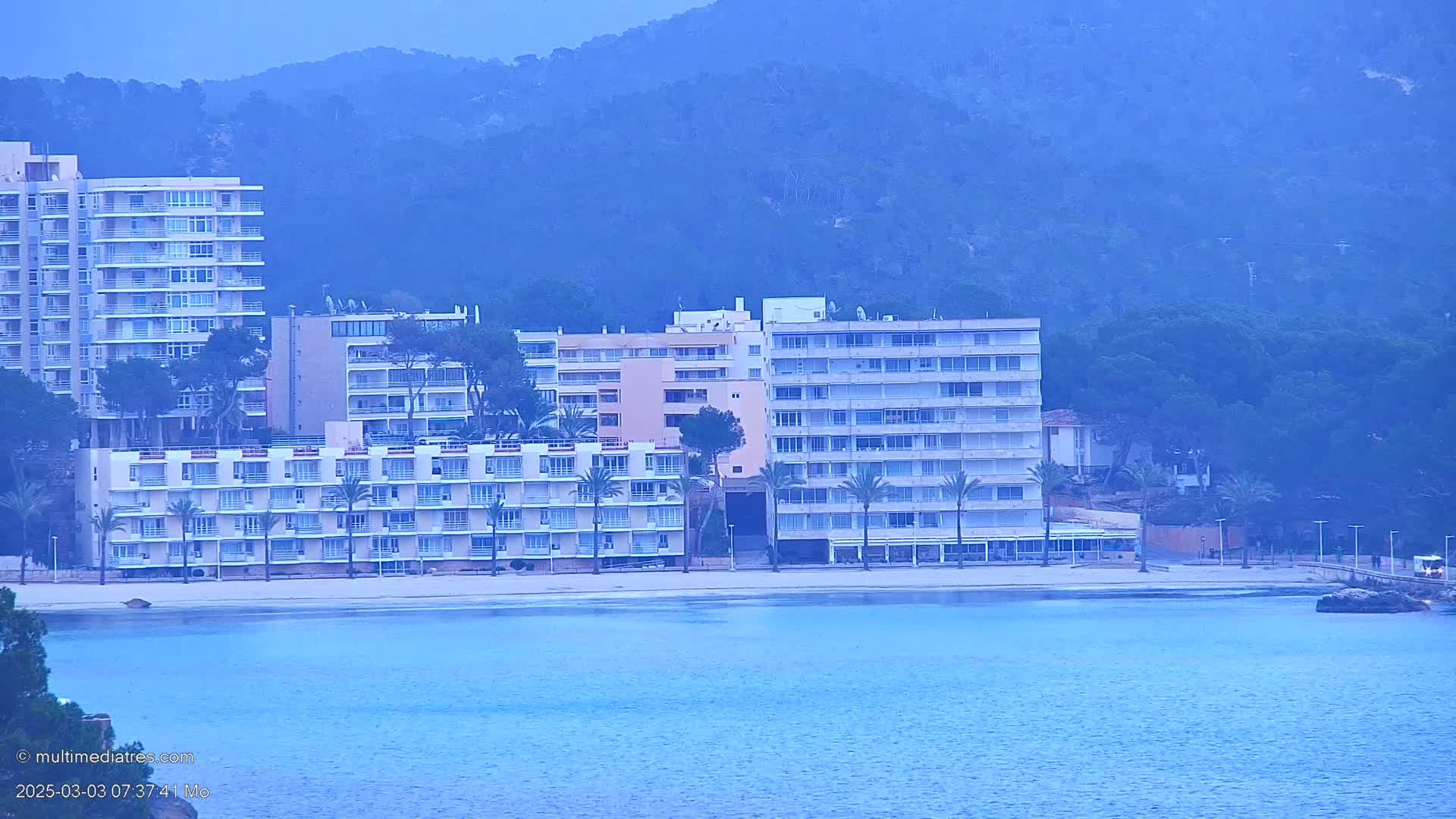 Several multi-story buildings line a sandy beach next to calm blue water, with hazy mountains in the background under an overcast sky.