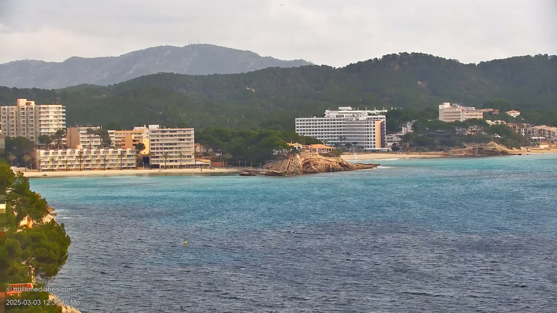 A coastal scene shows several hotels and buildings along a sandy beach, with a small rocky island in the turquoise water under an overcast sky.