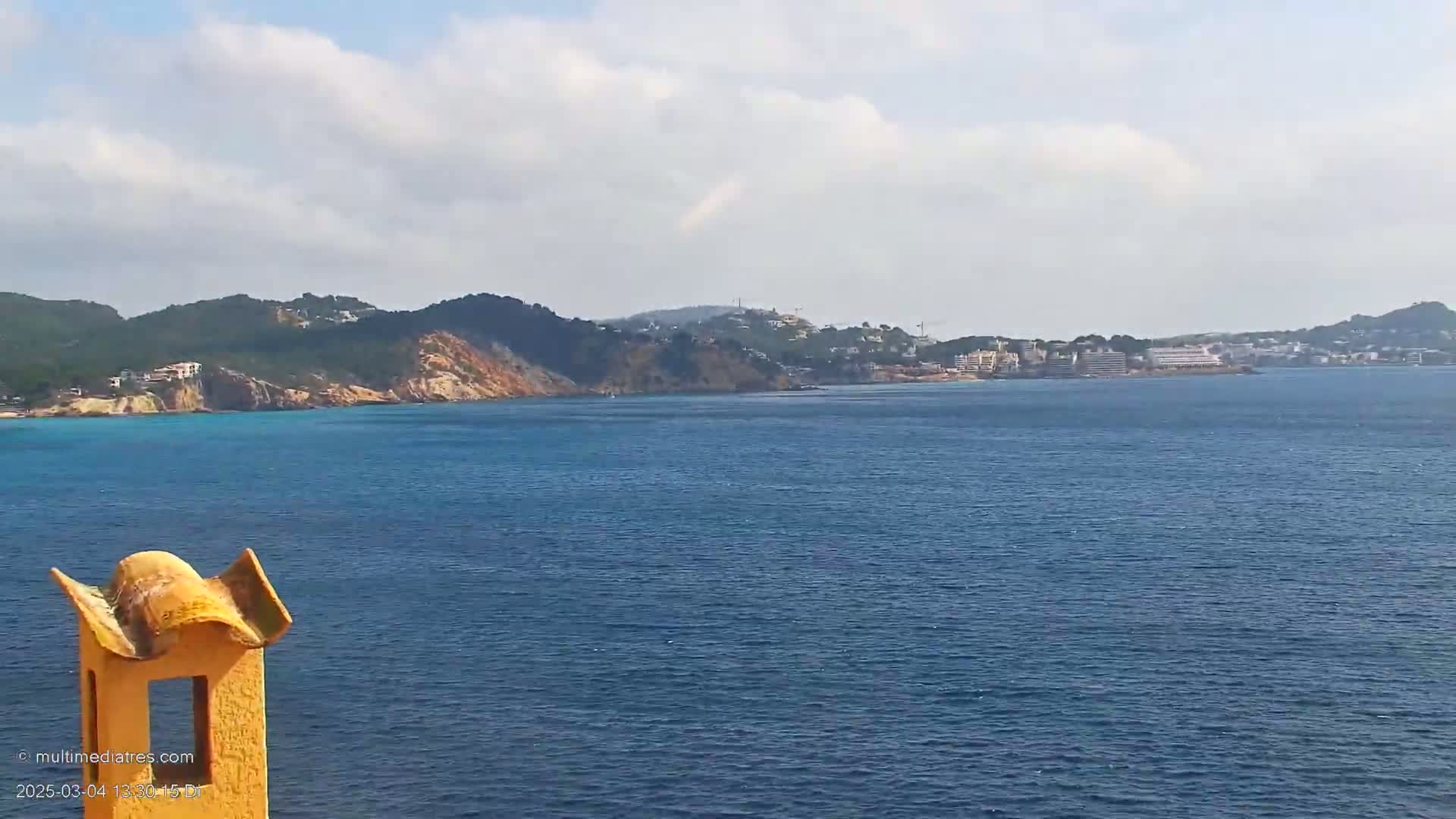 A partly cloudy day reveals a coastal scene of blue water, a distant town nestled against green hills, and a portion of a yellow structure in the foreground.