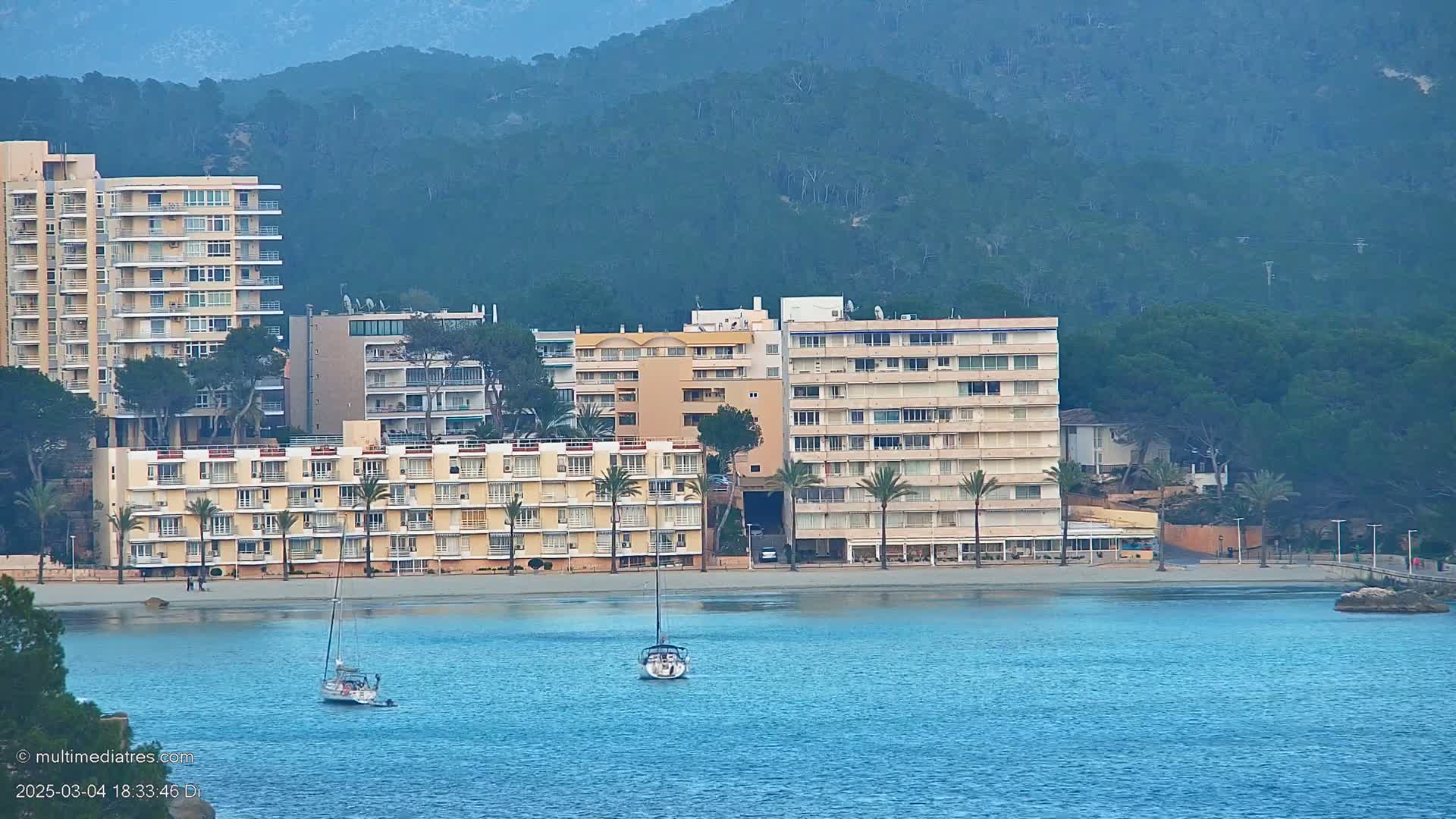 Multi-story buildings line a sandy beach, with sailboats in calm blue water and a mountain covered in dark green trees in the background.