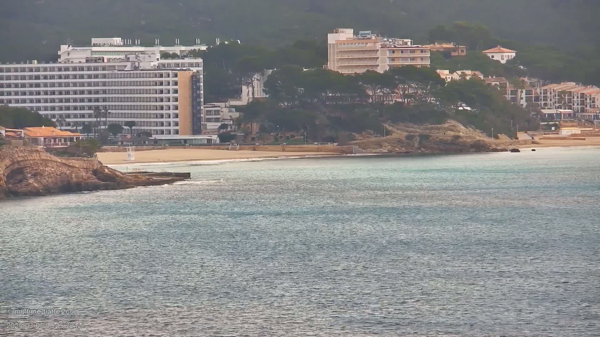 A sandy beach with several hotels and buildings behind it is seen from across calm water under an overcast sky.