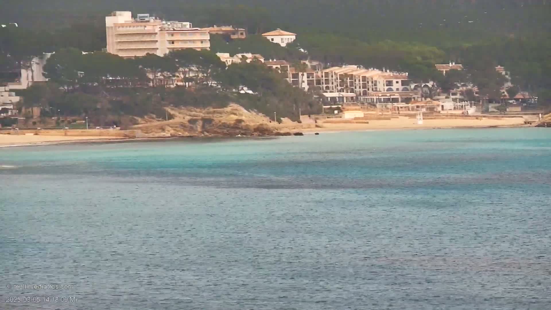 A sandy beach with turquoise water stretches out in front of buildings nestled on a hillside, under an overcast sky.