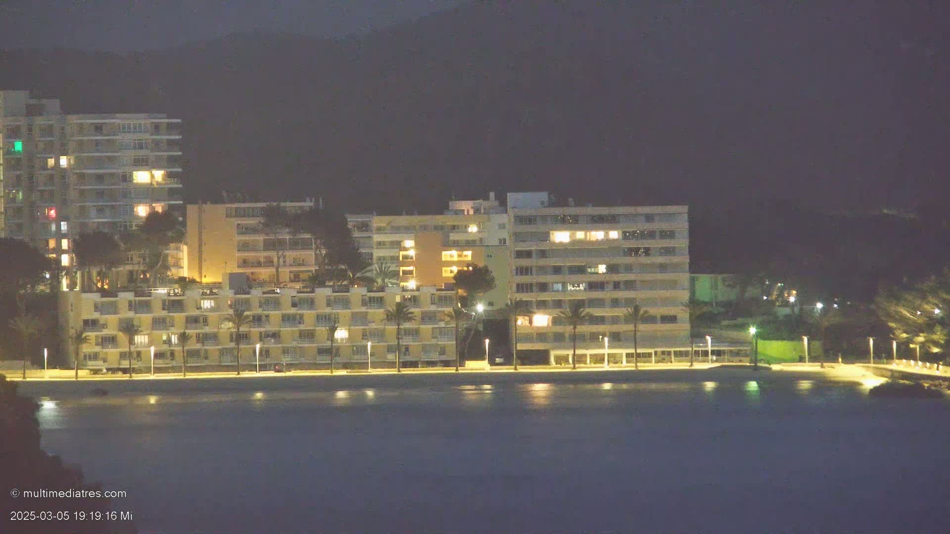 At night, several illuminated buildings line a calm body of water, with palm trees and hills in the background.