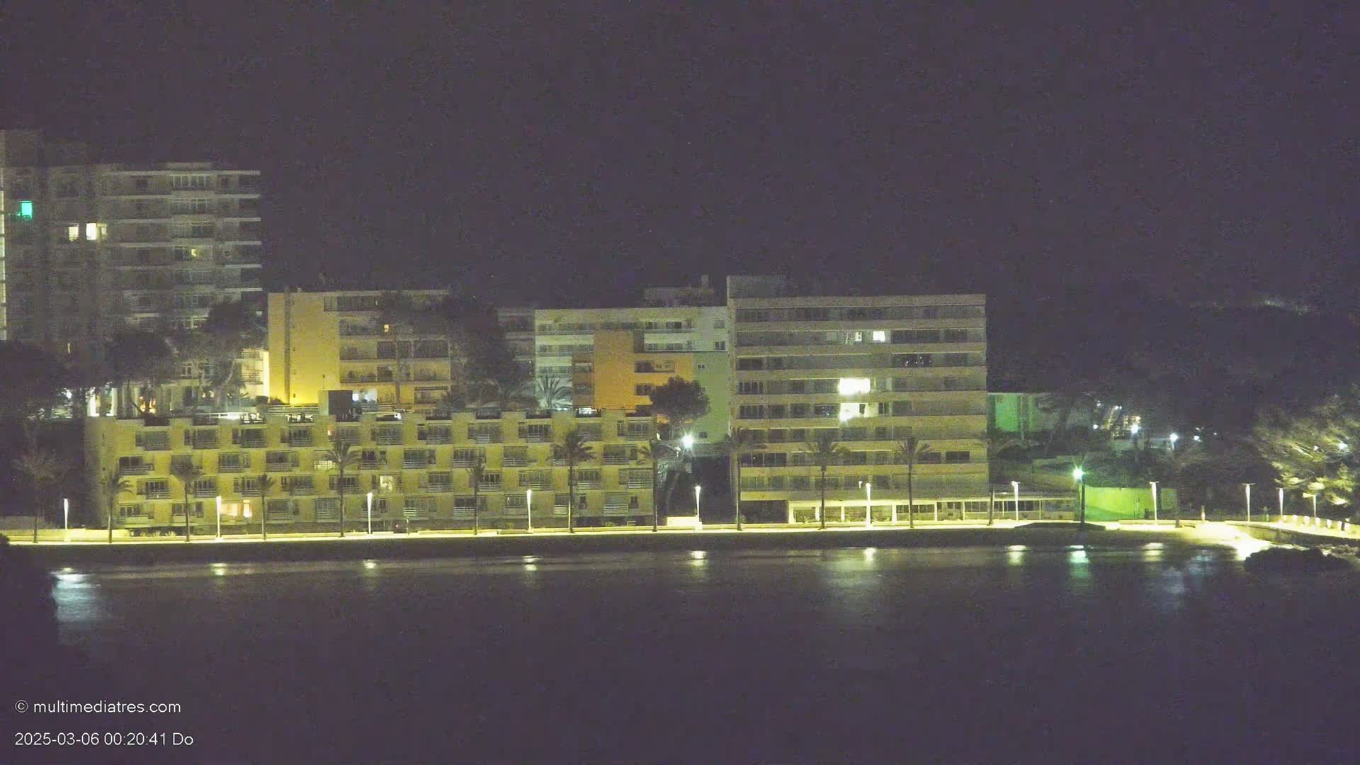 A row of illuminated multi-story buildings sits along a dark body of water at night.