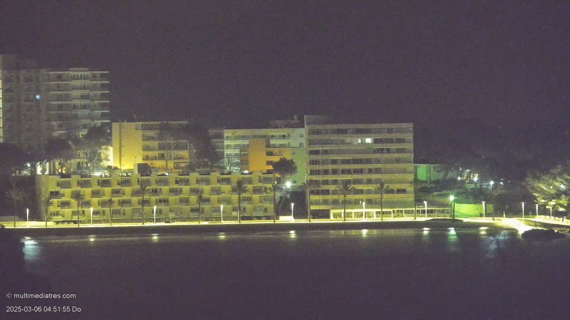 A nighttime view of several illuminated multi-story buildings situated along a waterfront, with palm trees lining the shore.