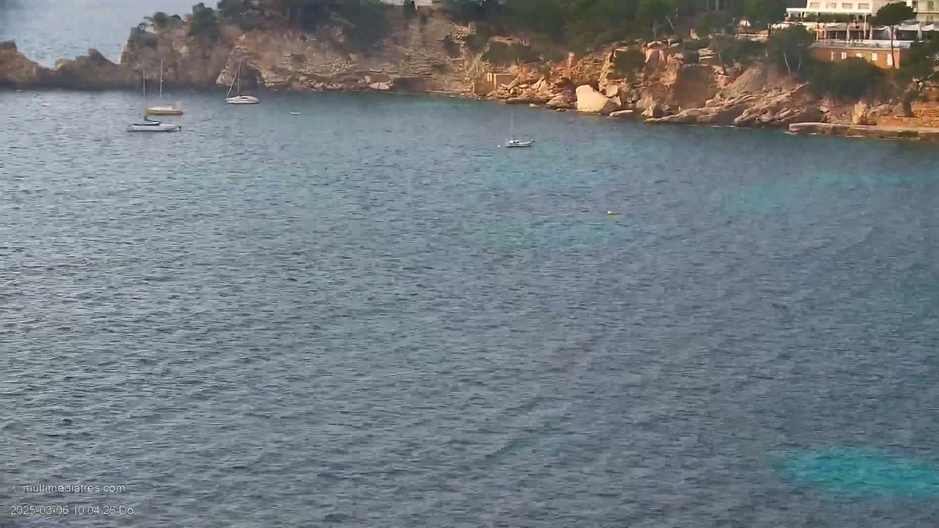 Several sailboats are anchored in a calm, blue-green bay next to a rocky, vegetated coastline.