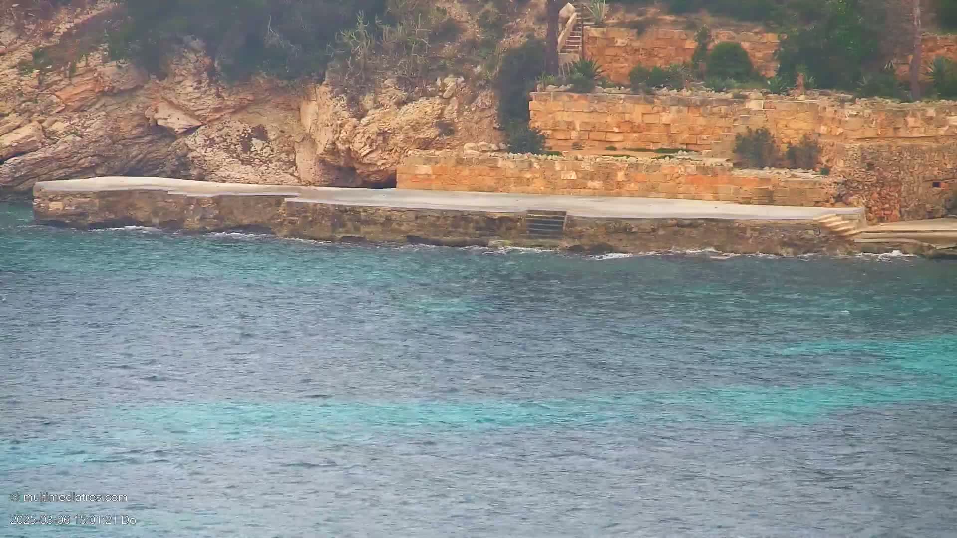 A cement platform extends from a rocky, tan-colored cliff into a calm, teal-colored sea.
