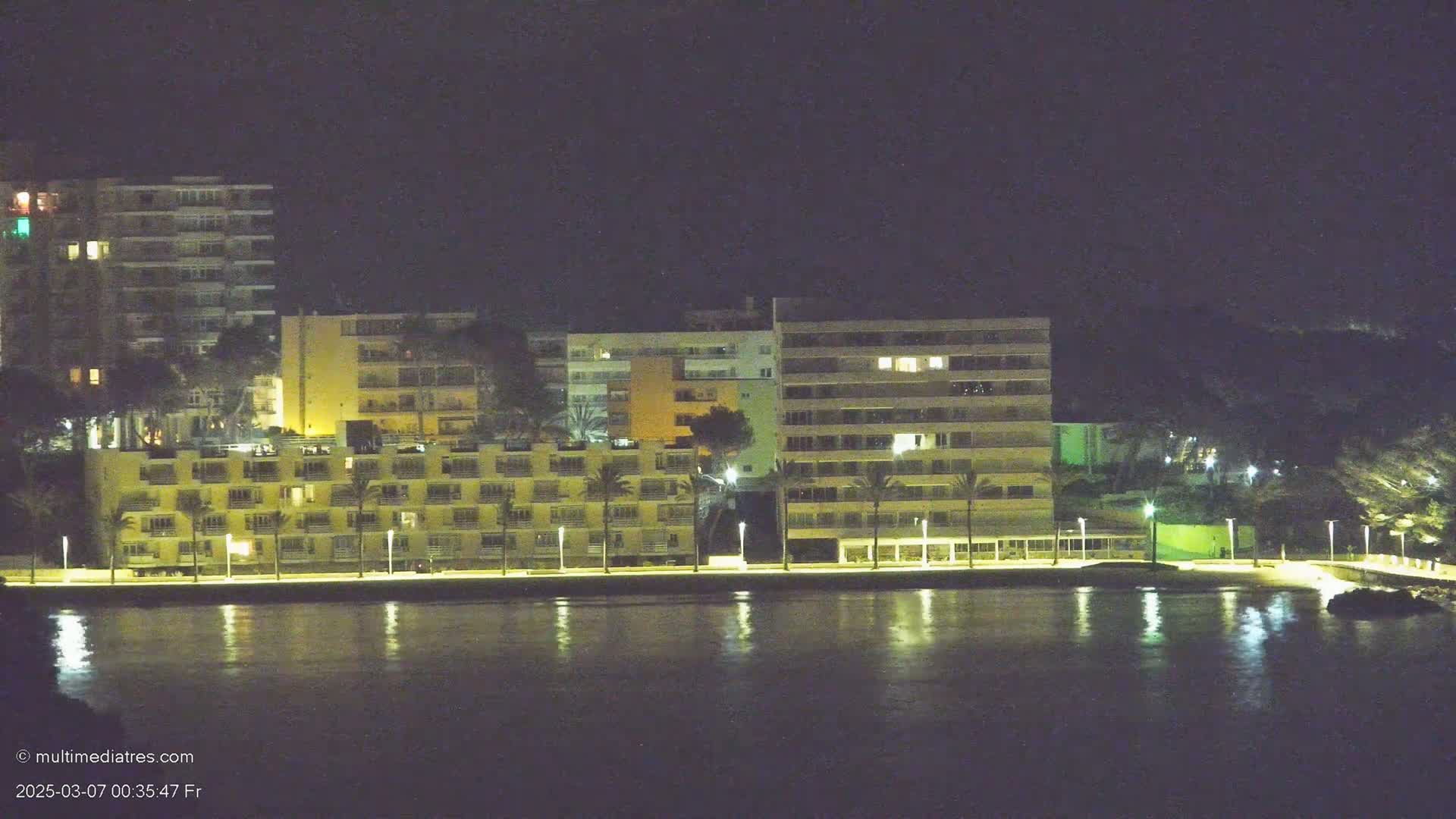 Illuminated apartment buildings line a waterfront at night, their lights reflecting on the calm water.