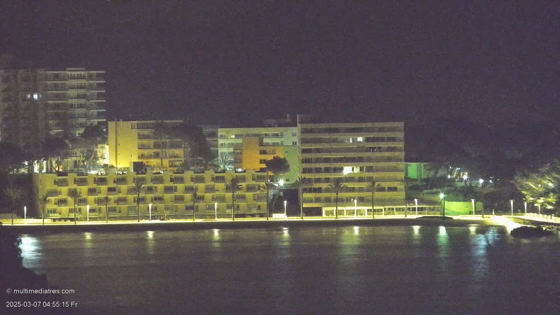 A line of illuminated multi-story buildings sits along a calm body of water at night.