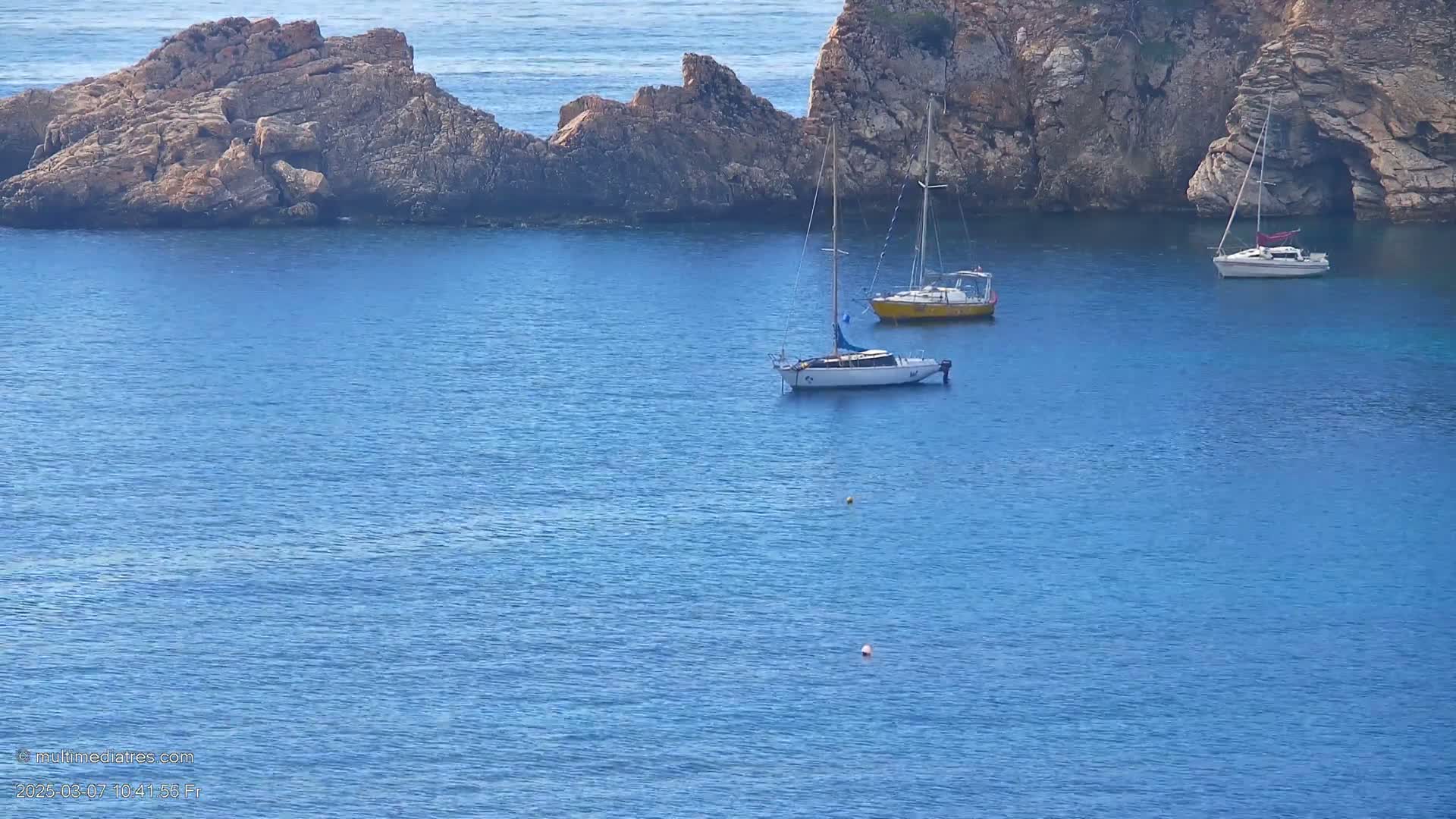 Three sailboats are anchored in a calm, blue sea near rocky cliffs.