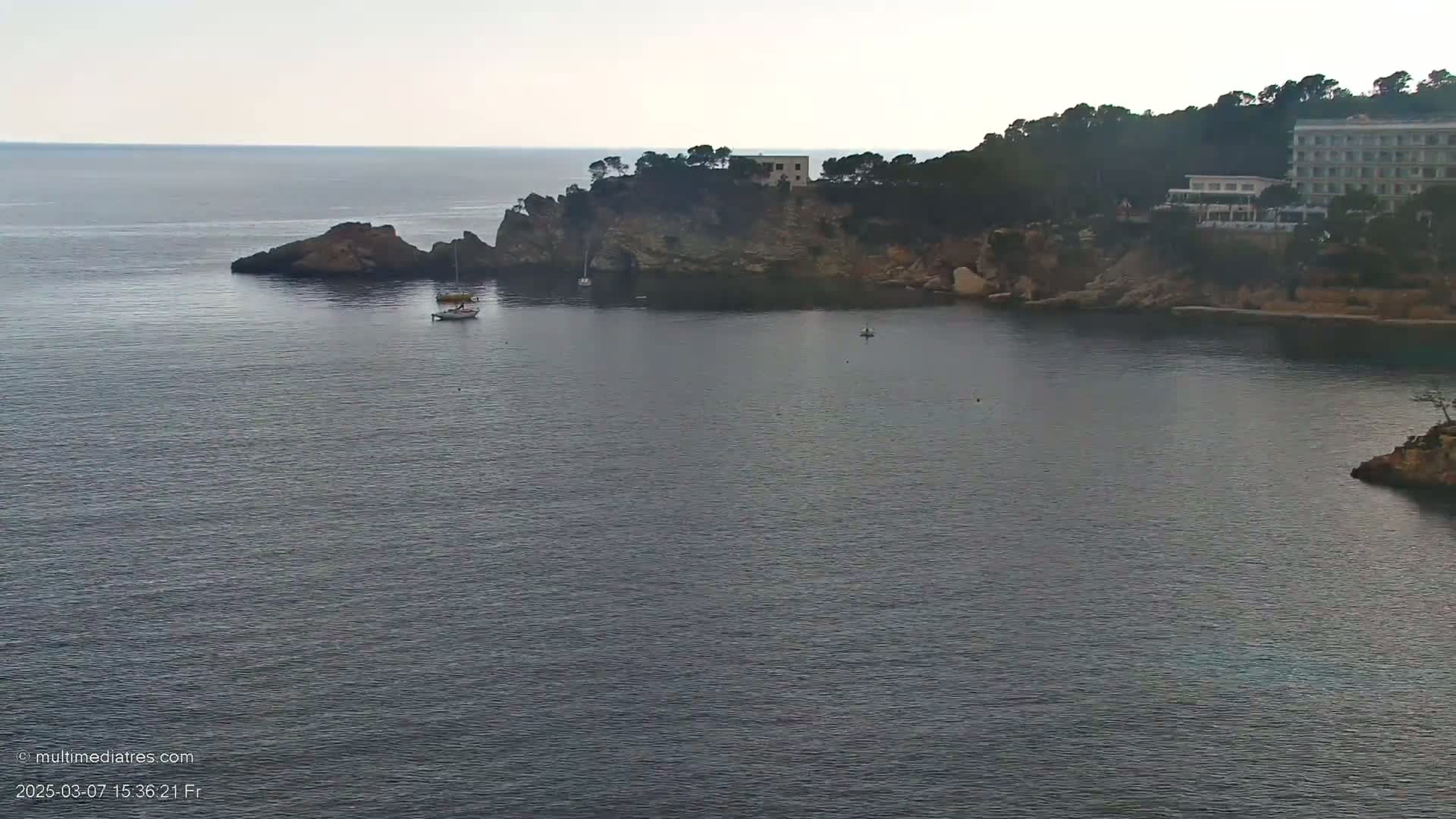 A calm, gray-toned sea surrounds a rocky cove with a building nestled among trees on the shore.