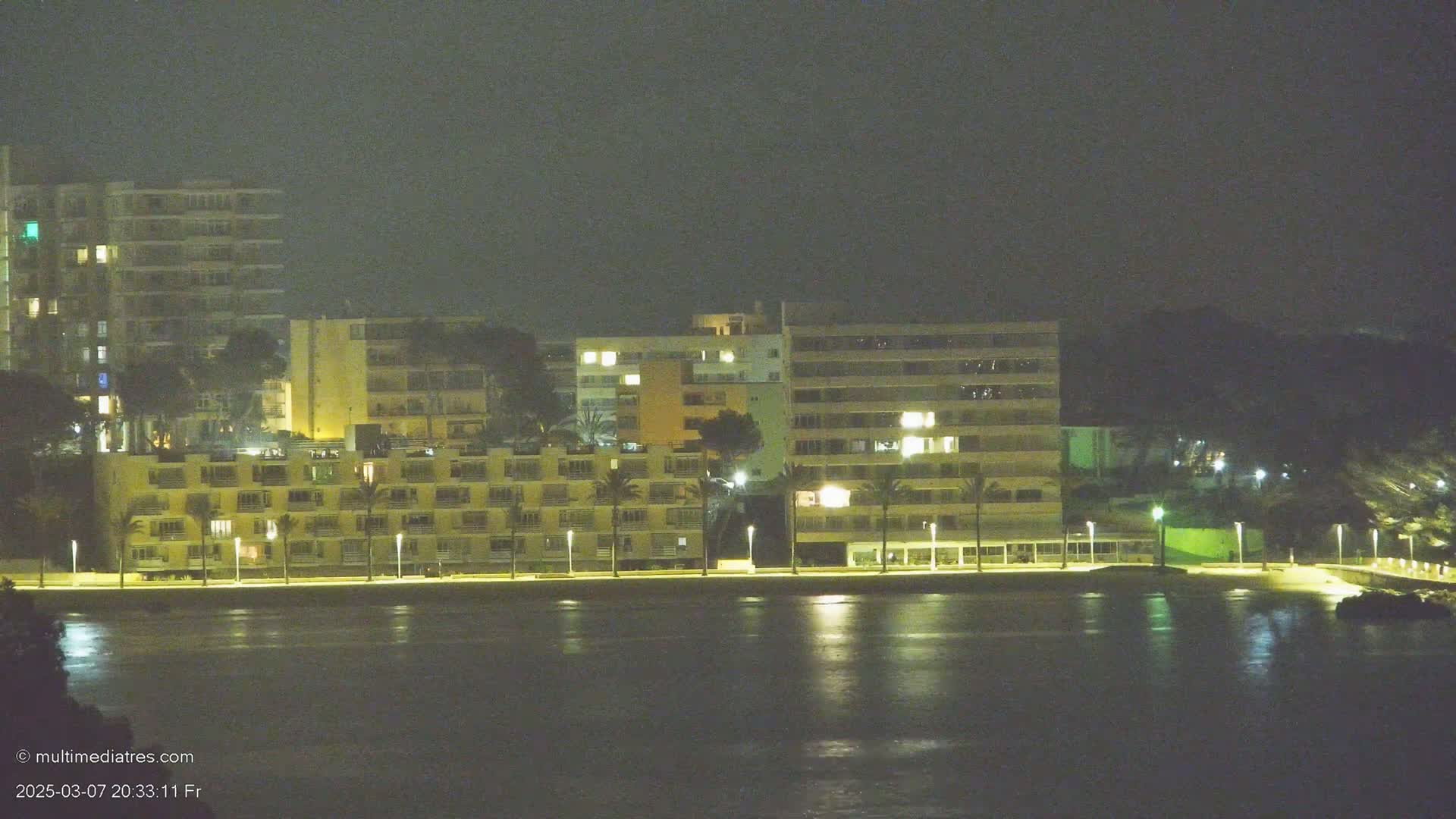 A nighttime view of several multi-story buildings illuminated along a waterfront, with calm water reflecting the city lights.