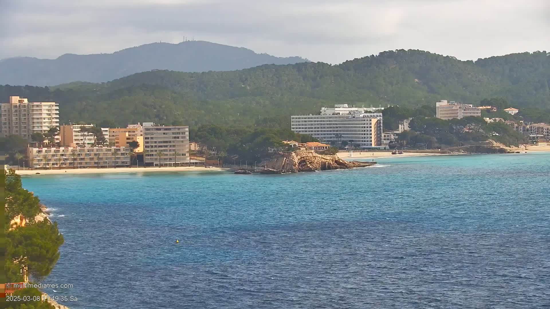 A picturesque coastal scene features several hotels and buildings situated along a sandy beach with turquoise water, backed by lush green hills under a partly cloudy sky.