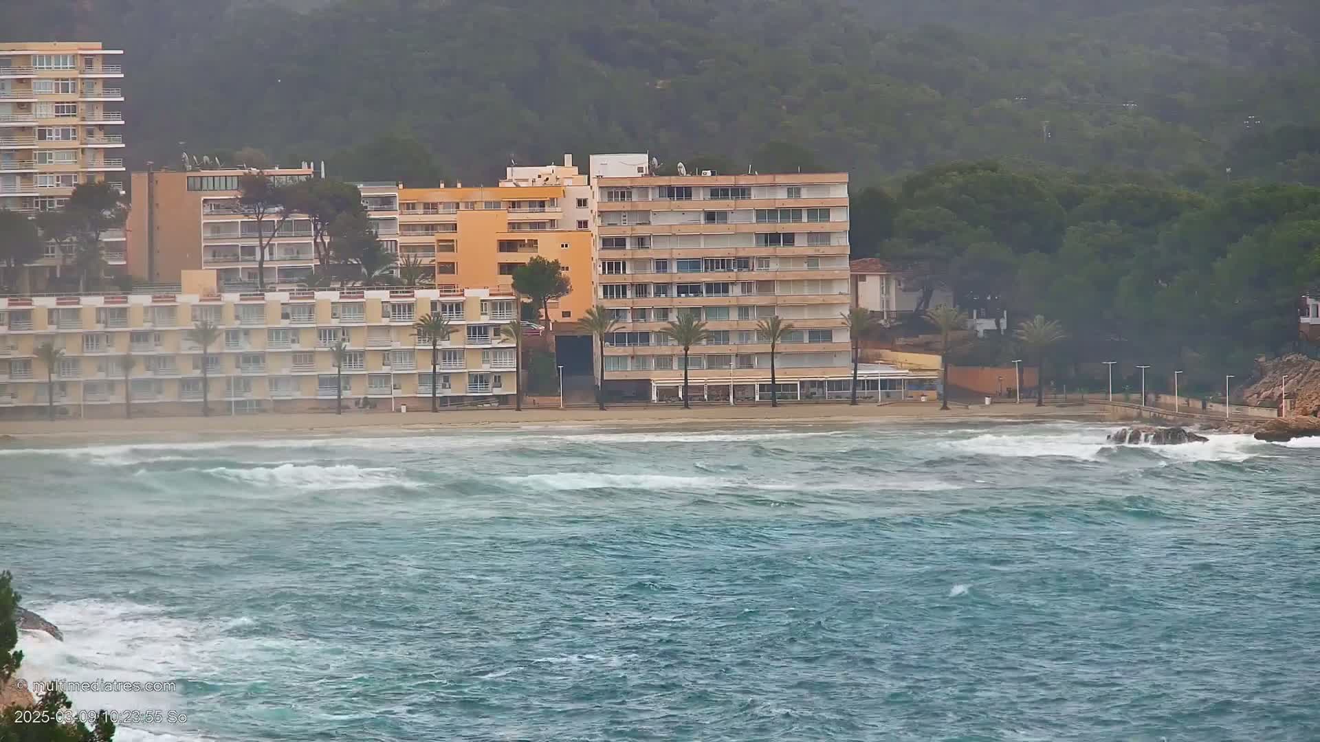 A sandy beach with several multi-story buildings is lashed by choppy ocean waves under an overcast sky.