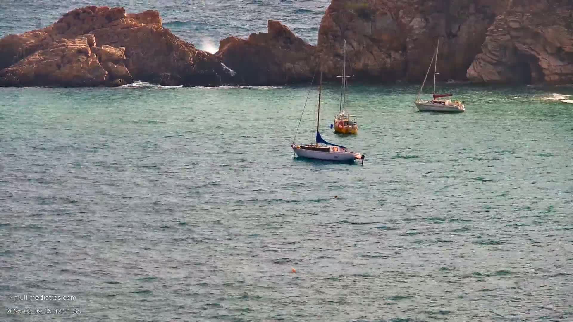 Three sailboats are anchored in calm, clear water near rocky outcrops.