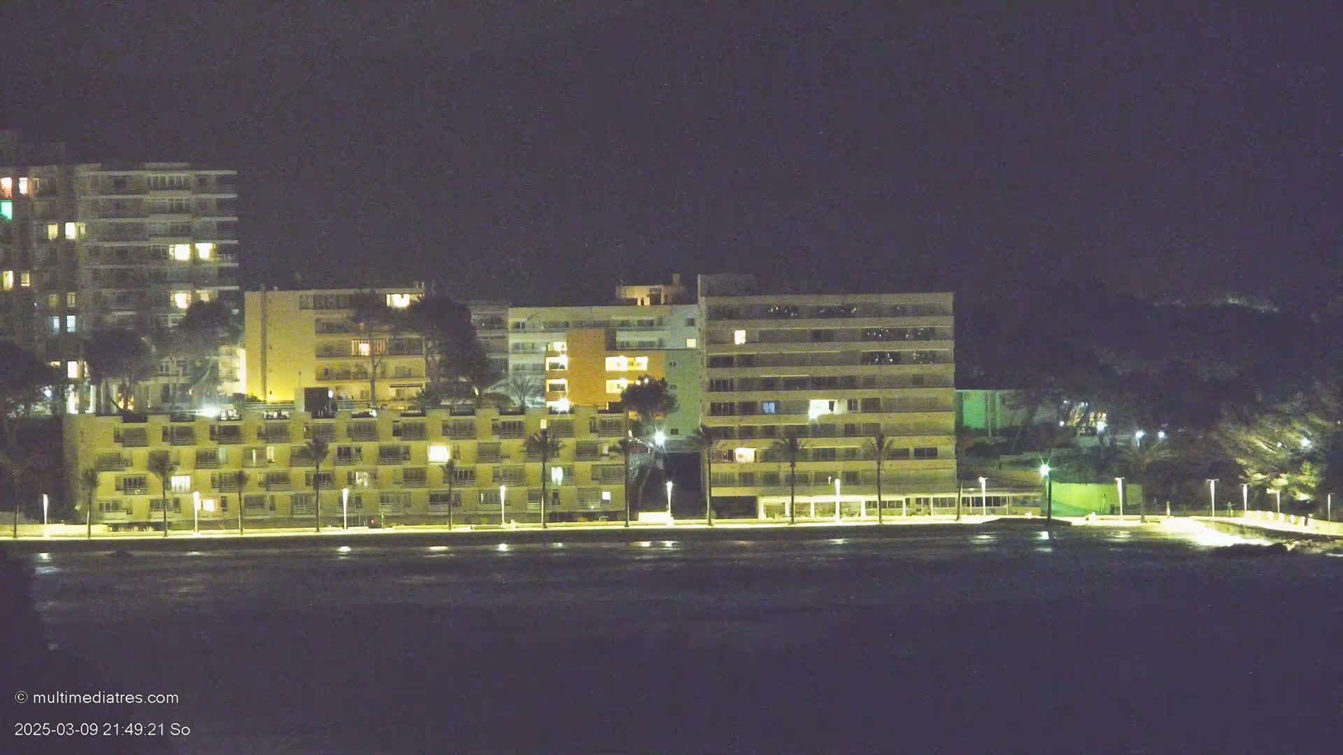 A row of illuminated apartment buildings lines a dark, calm body of water at night.