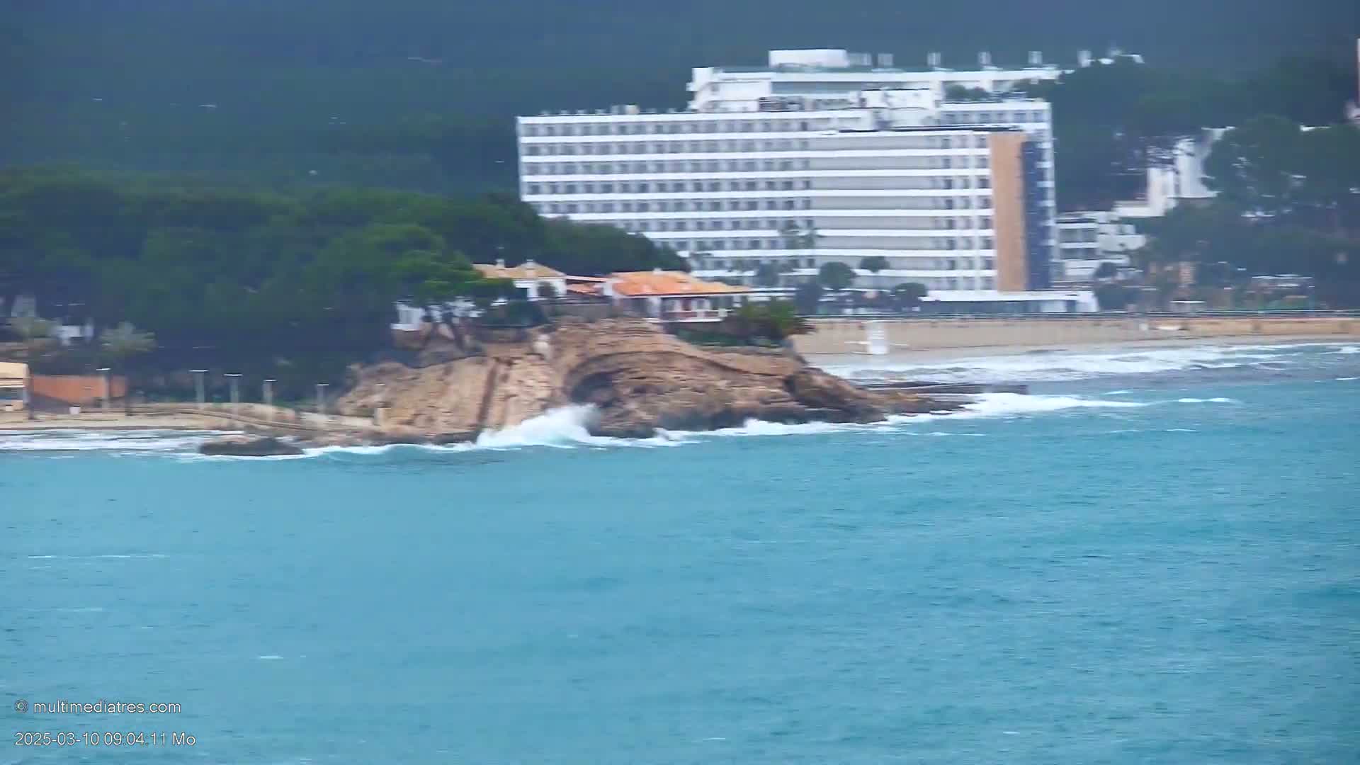 Waves crash against a rocky outcrop near a beach with a large hotel and other buildings in the background on an overcast day.