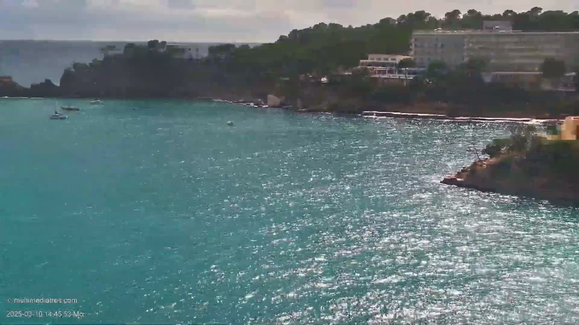 A teal-colored body of water with small waves reflects sunlight, with a few small boats visible in the distance near a rocky coastline and a large building on a hillside under a partly cloudy sky.