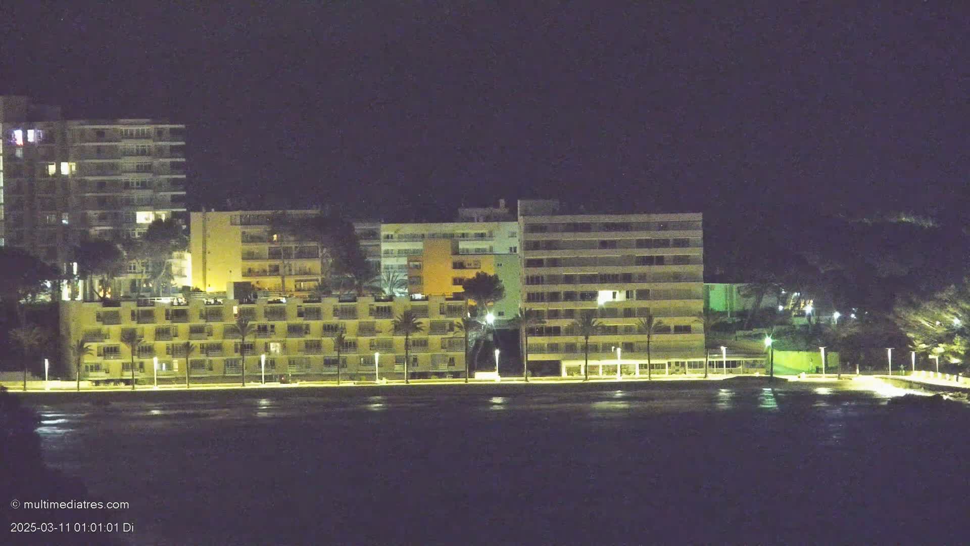 A row of illuminated apartment buildings stands along a dark waterfront at night.