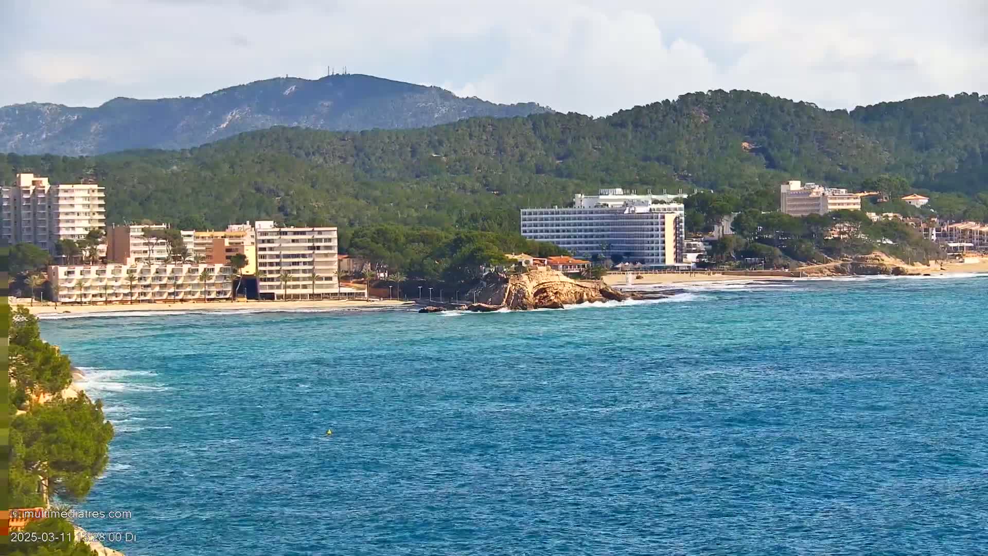 A partly cloudy day shows a beach with several large hotels and a rocky outcrop in the turquoise water, backed by green hills and mountains.