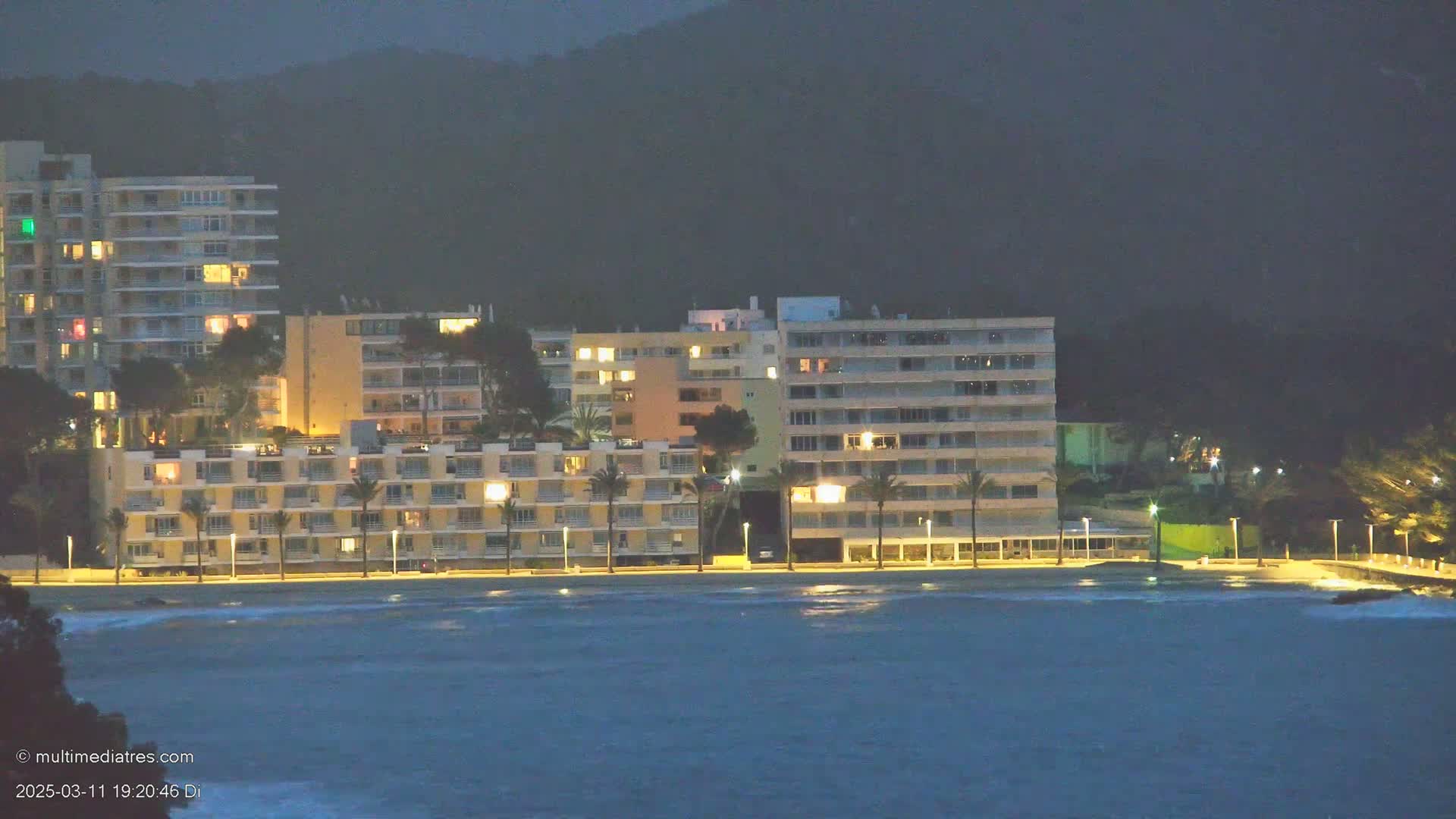 A row of lit multi-story buildings lines a beach at night, under a dark, possibly stormy sky, with a somewhat choppy sea in the foreground.