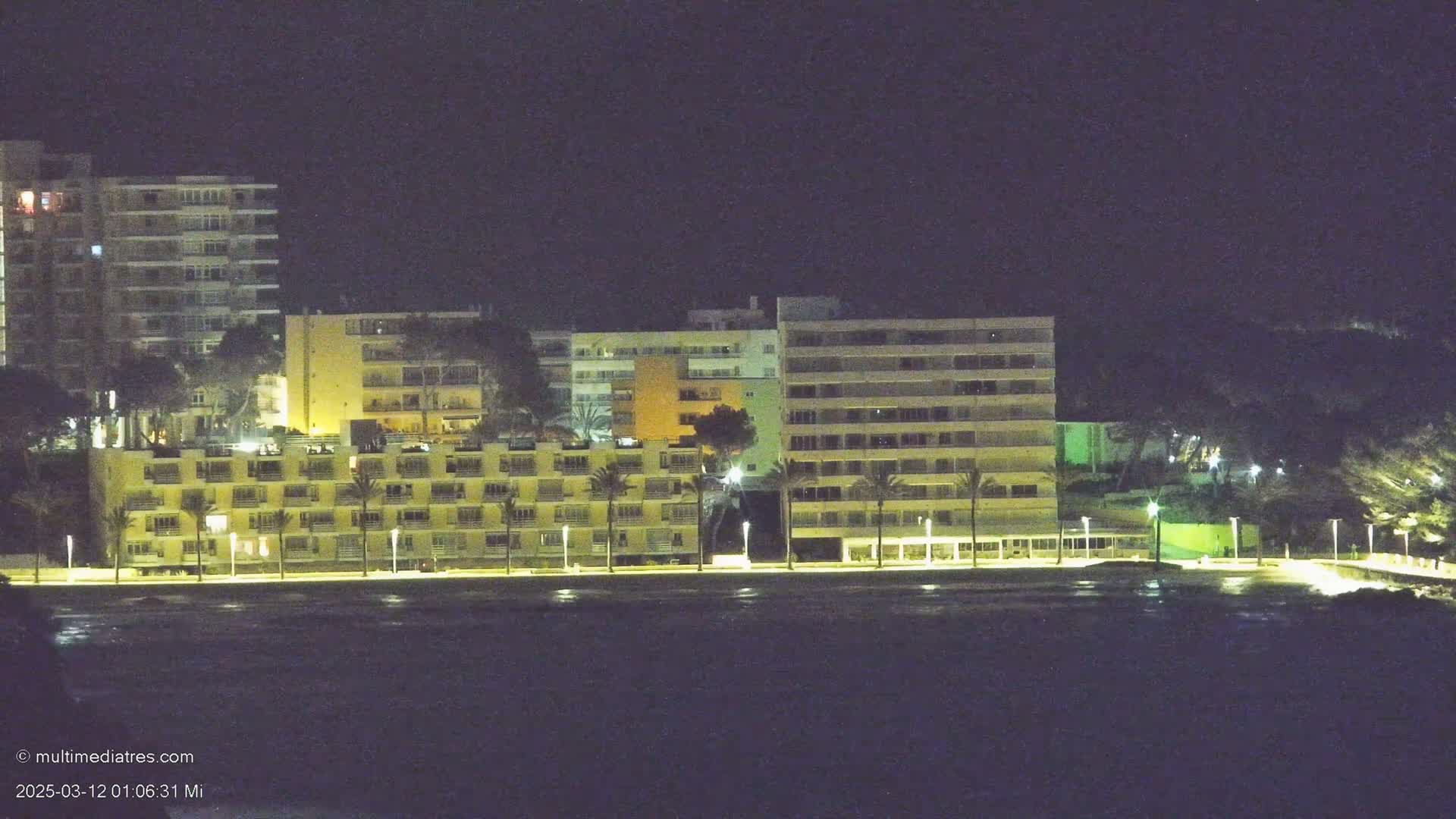 A row of illuminated multi-story buildings sits along a dark body of water at night.