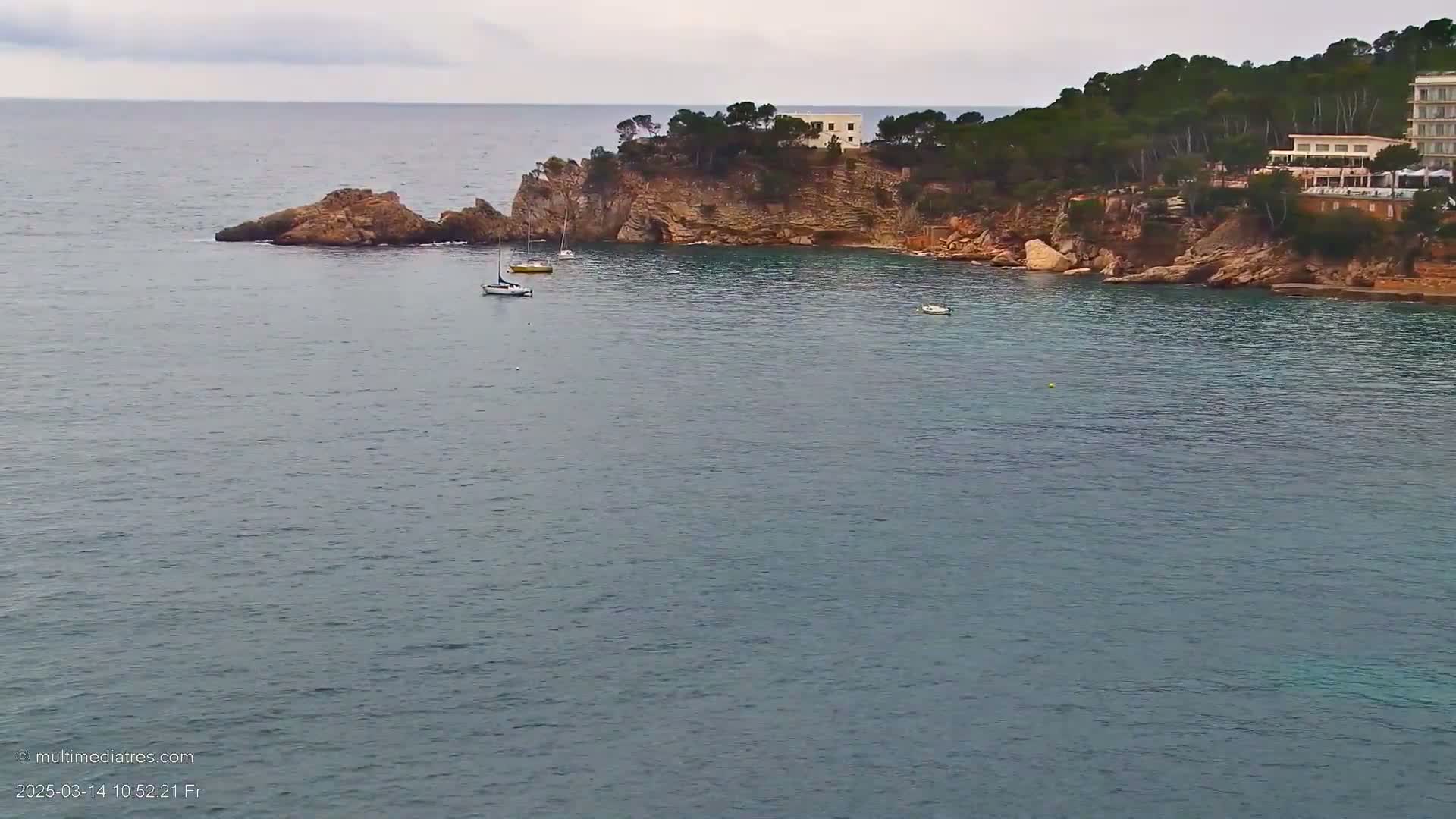 A rocky coastline with a few small boats in calm, gray water under an overcast sky.