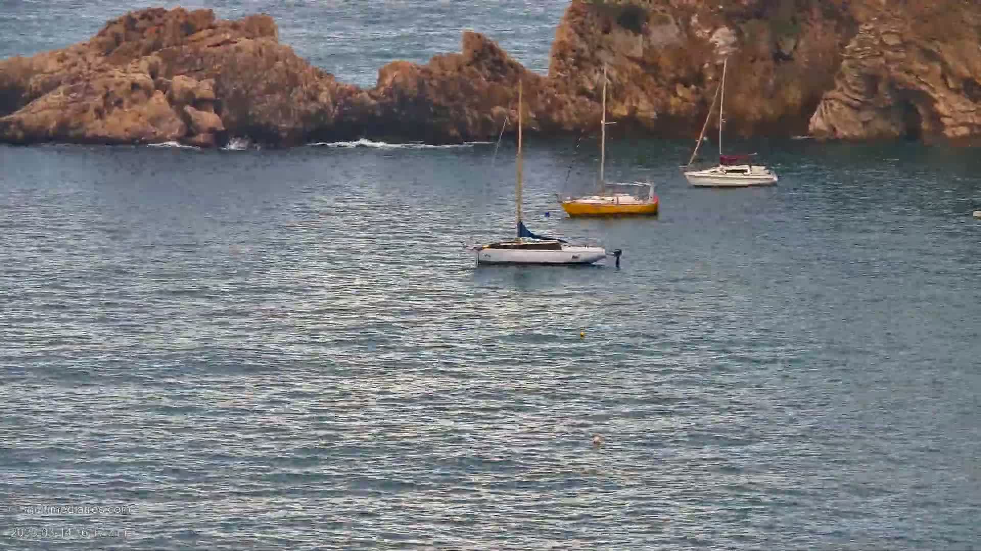 Three sailboats are anchored in a calm sea near rocky outcrops.