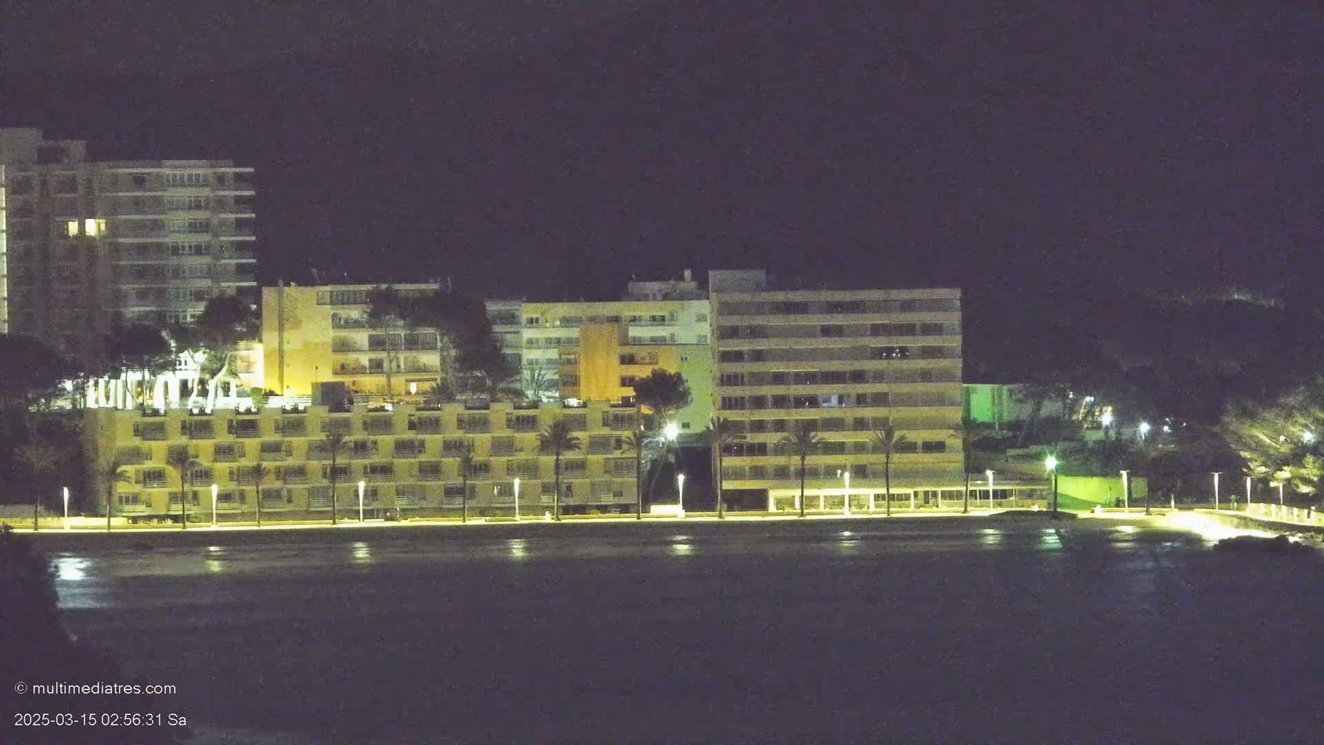 At night, several illuminated multi-story buildings line a dark, calm body of water.