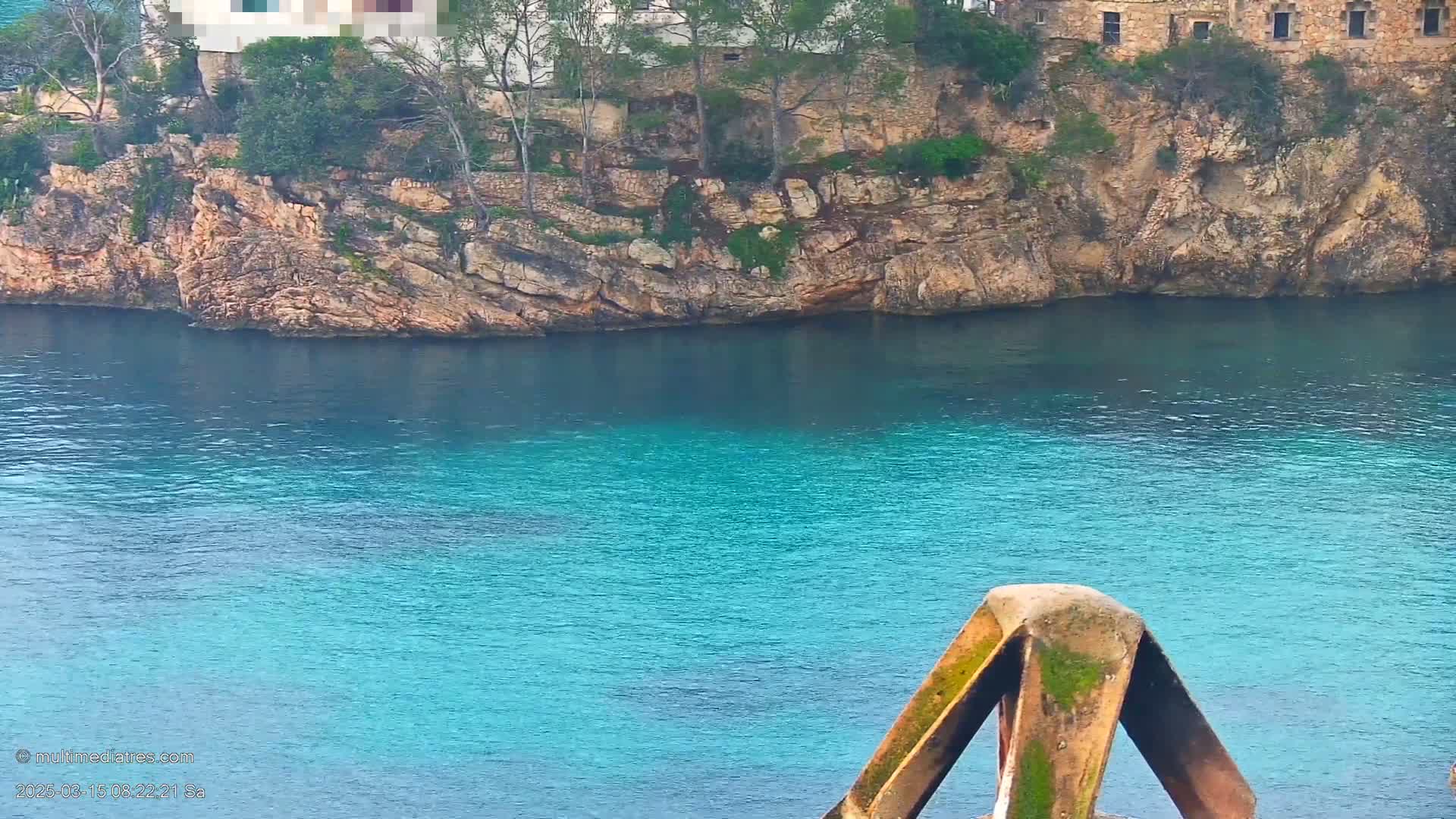 A rocky cliff with sparse vegetation overlooks a calm, turquoise body of water, partially obscuring a building on the clifftop.